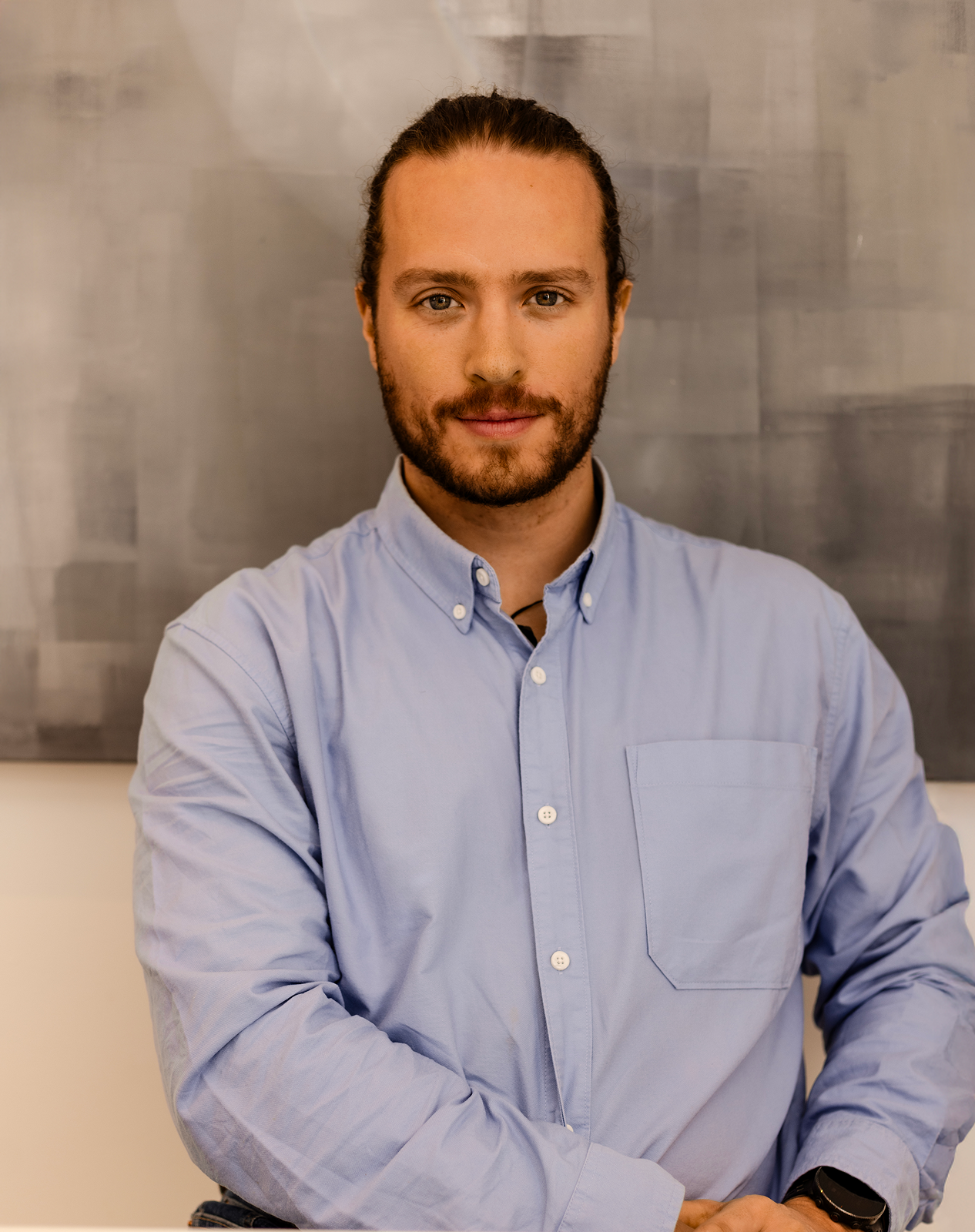 Portrait of a executive coach Greg Chambers looking directly at the camera. Beard and long hair, wearing a light blue button-down shirt against a grey background.