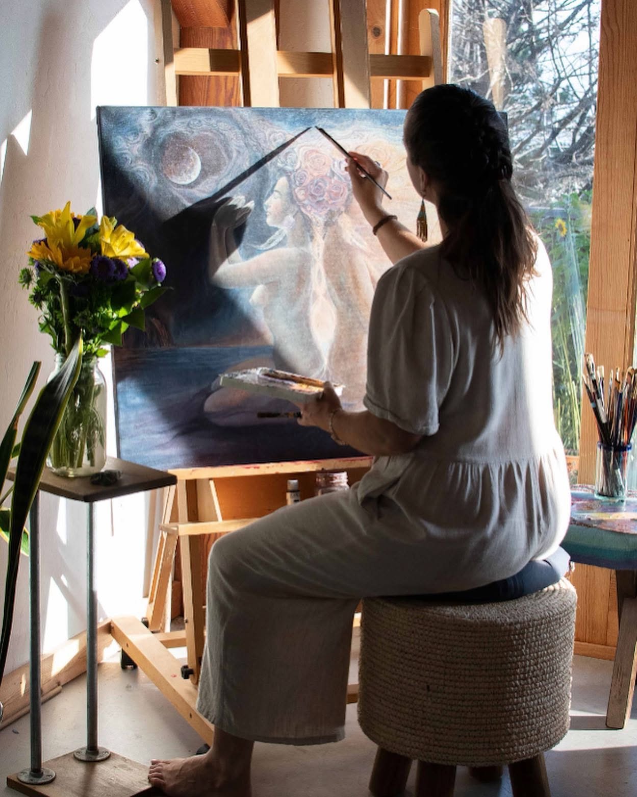 A woman in a light-colored dress is sitting on a round stool, painting a surreal scene of two female figures with floral hair and a moon in the sky on a large canvas. She is holding a palette in one hand and a paintbrush in the other, working indoors near a window, with sunlight coming in. There is a vase of flowers on a small table to her side.