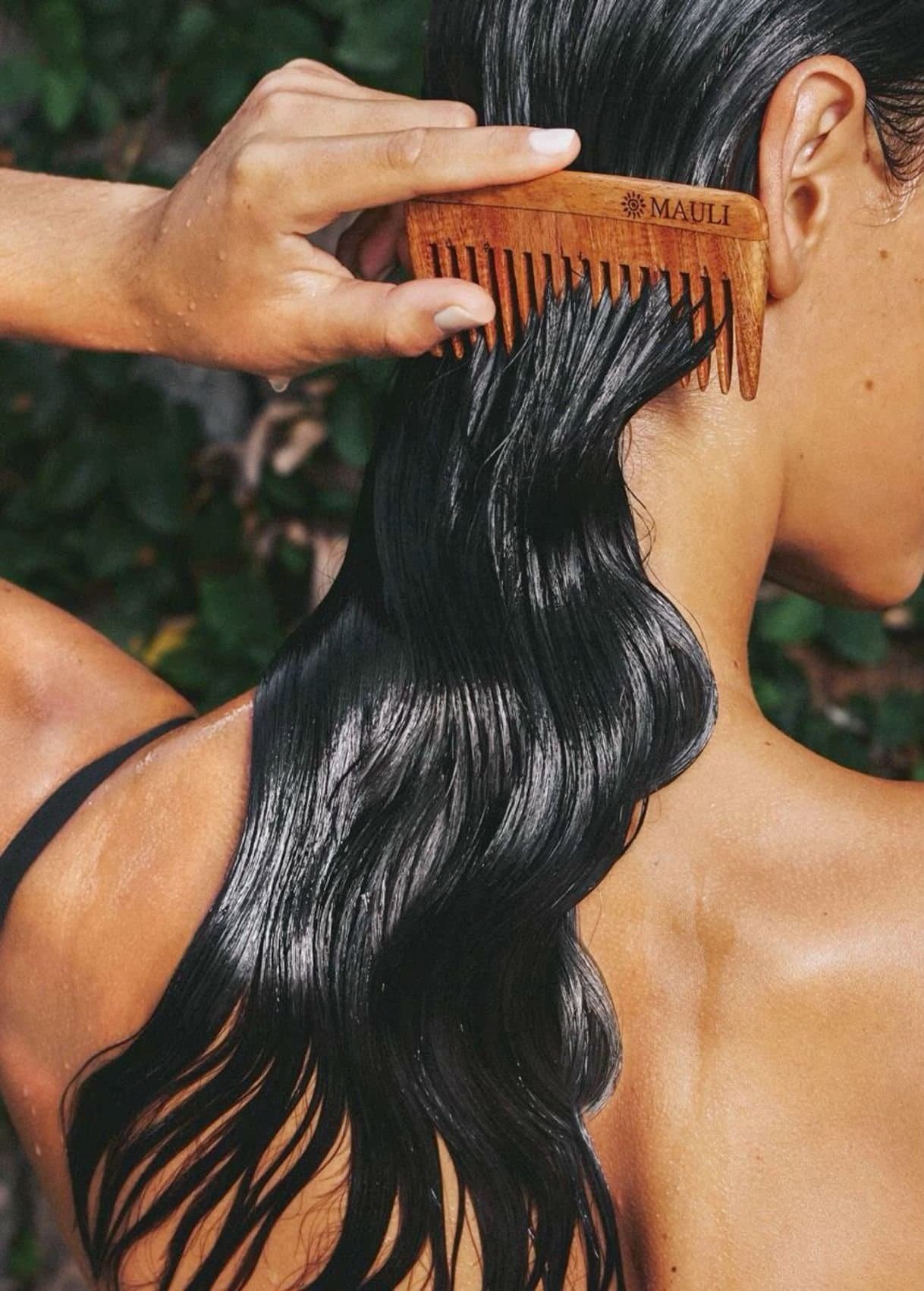 Close-up of a person with wet, dark hair using a wooden comb to detangle or groom hair, outdoors with greenery in the background.