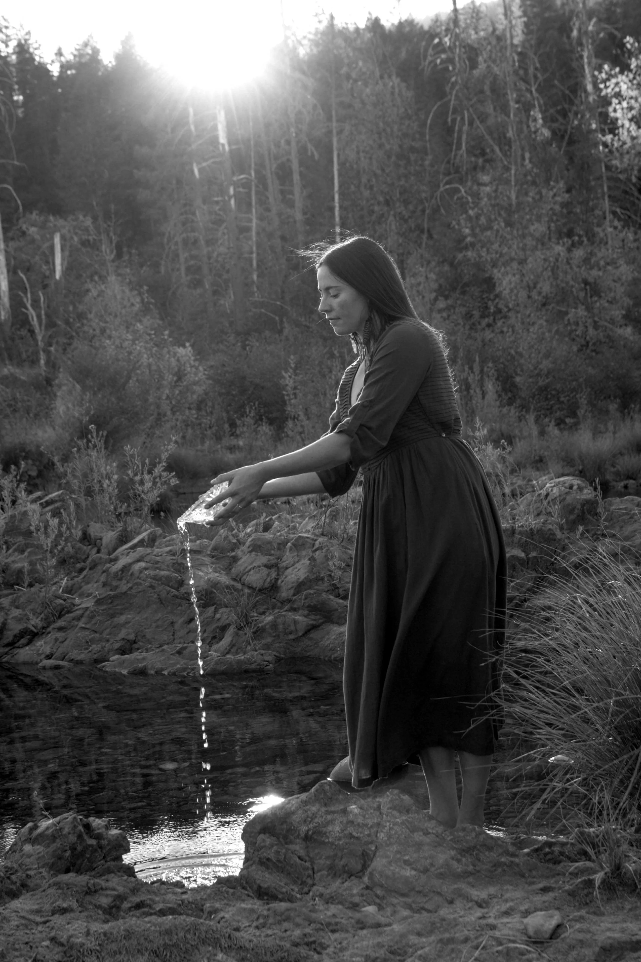 A woman in a dress with long dark hair standing in a creek, pouring water from a glass, with the sun setting behind a forested hillside.