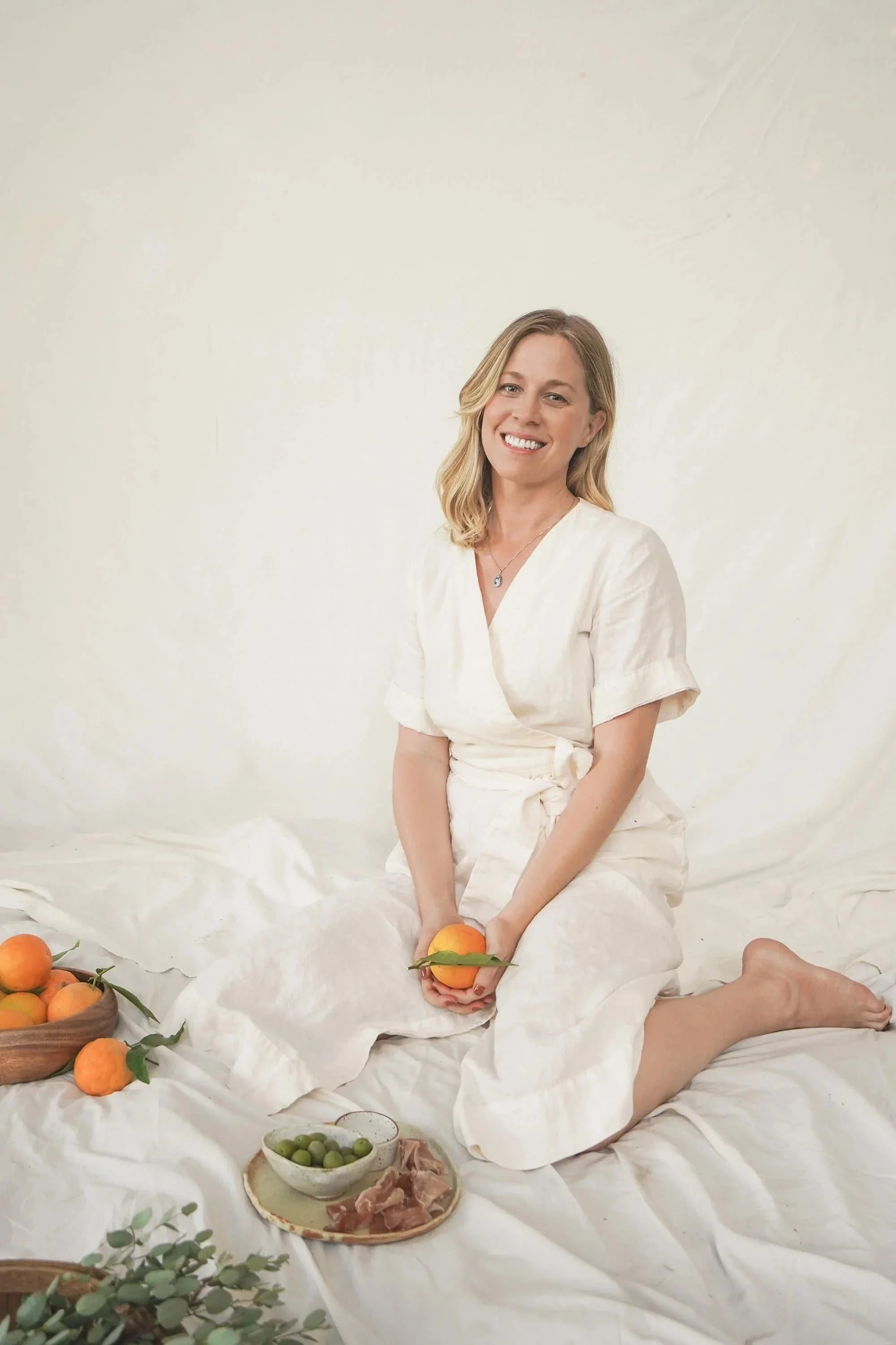 Female founder Christina Tidwell with blonde hair smiling and sitting on her knees surrounded by peaches, a bowl of green olives, and a plate of sliced meat, on a white fabric background.