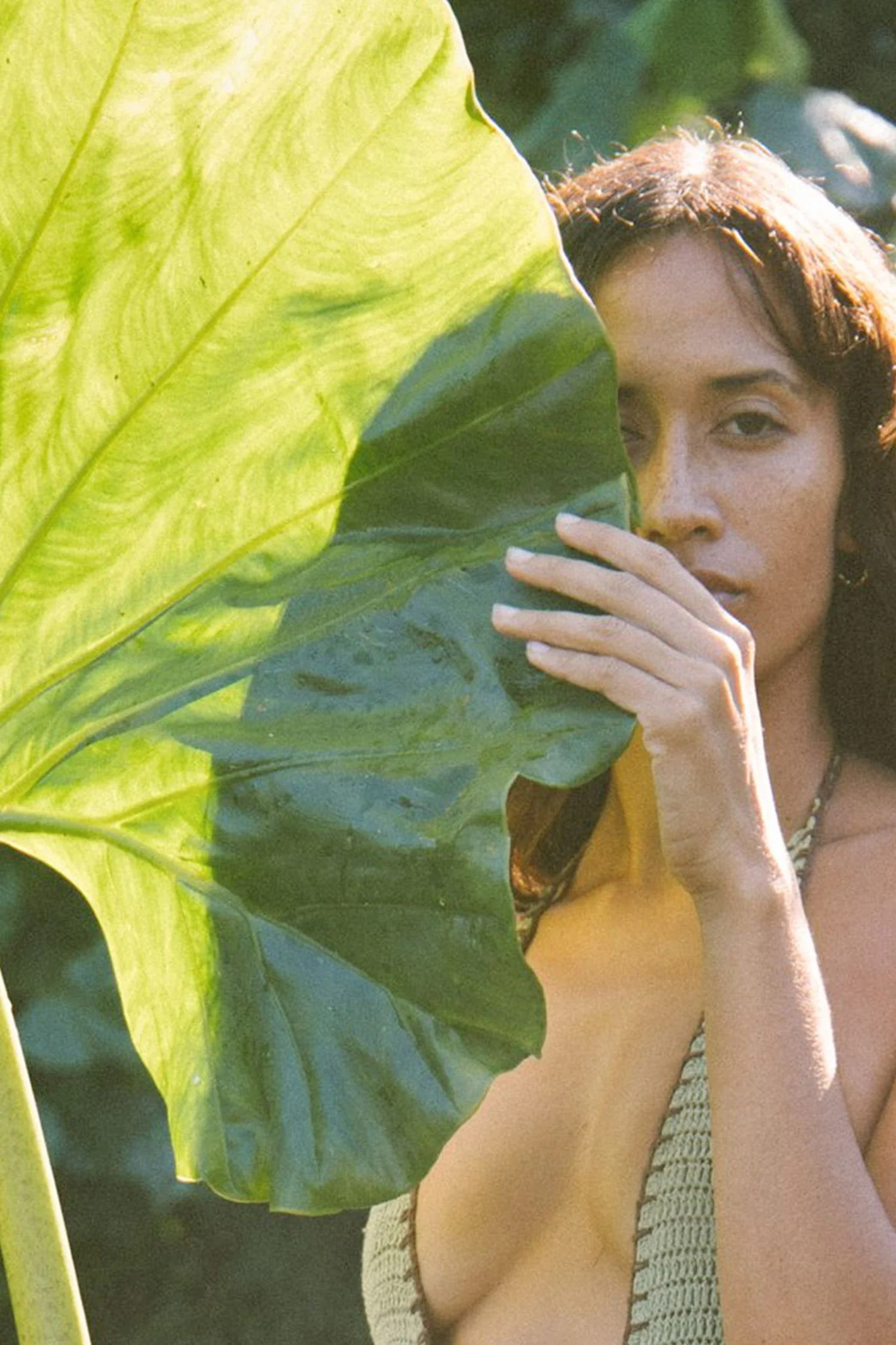 A woman with long brown hair holding a large green leaf in front of her face outdoors.