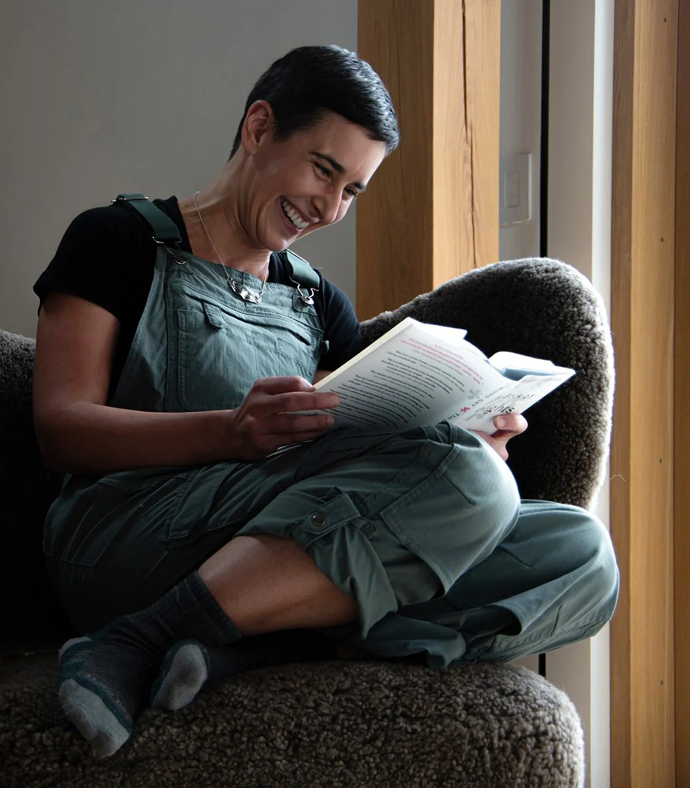 A woman with short dark hair is sitting on a cushioned chair, reading a book and smiling happily. She is wearing a black shirt, green overalls, and socks, with a necklace. The room has wooden walls and a window nearby, letting in natural light.