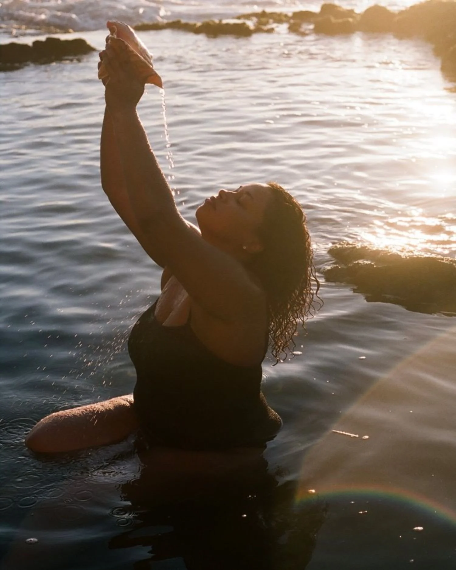 A woman with wet, curly hair is in the water at the beach, holding a conch shell up with water dripping from it during sunset.