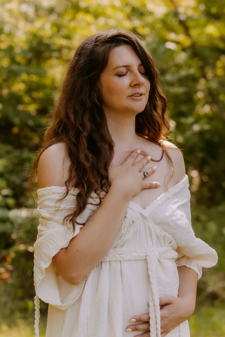 A woman with long, wavy brown hair stands outdoors with trees and sunlight in the background. She wears a loose, off-shoulder white dress and has her right hand on her chest and left hand on her stomach, with eyes closed and a serene expression.