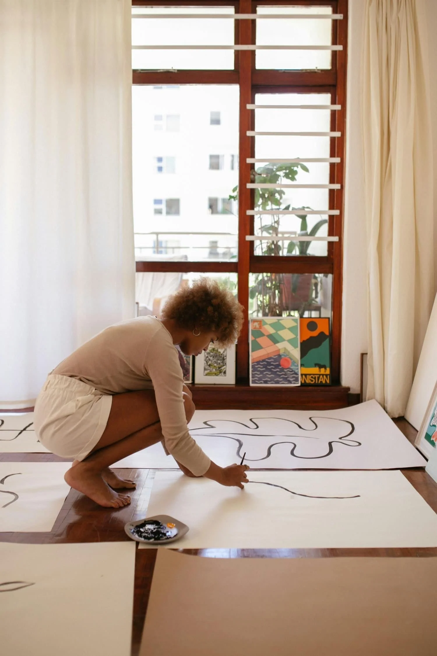 A woman with curly hair is crouching on the floor, painting a large abstract black line on a white sheet of paper or canvas.