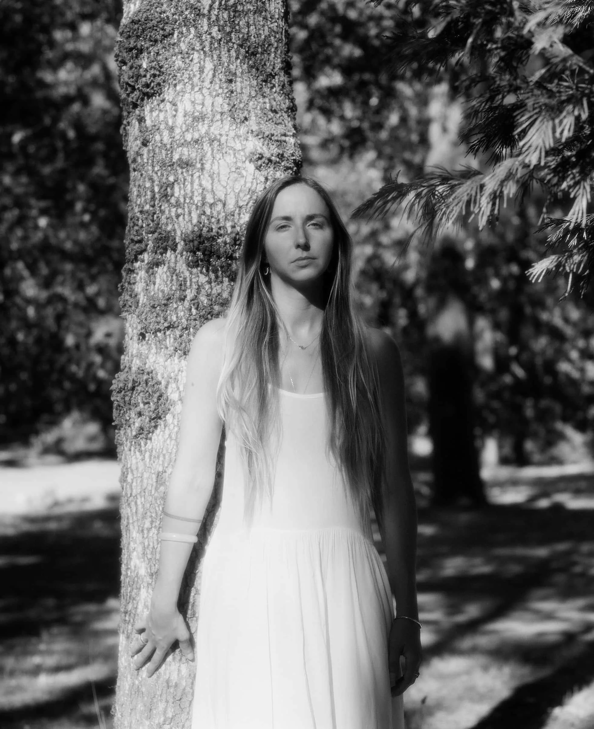 A woman with long hair wearing a sleeveless dress standing outdoors, leaning against a tree with her hand resting on the tree trunk, surrounded by blurred trees and foliage in the background for Le Petit 3.