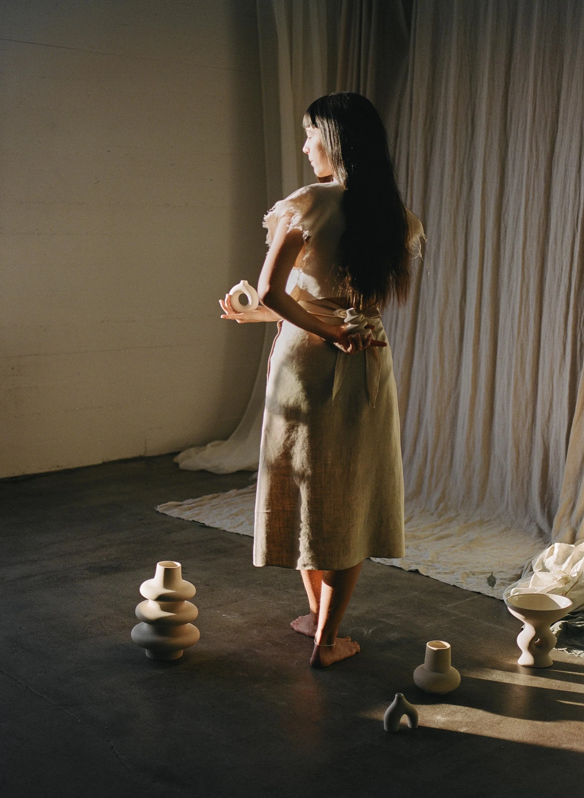 A woman standing barefoot in a room with neutral-colored walls and curtains, holding a small white sculpture. There are various white ceramic vases and bowls placed on the floor around her, and a large fabric backdrop behind her.