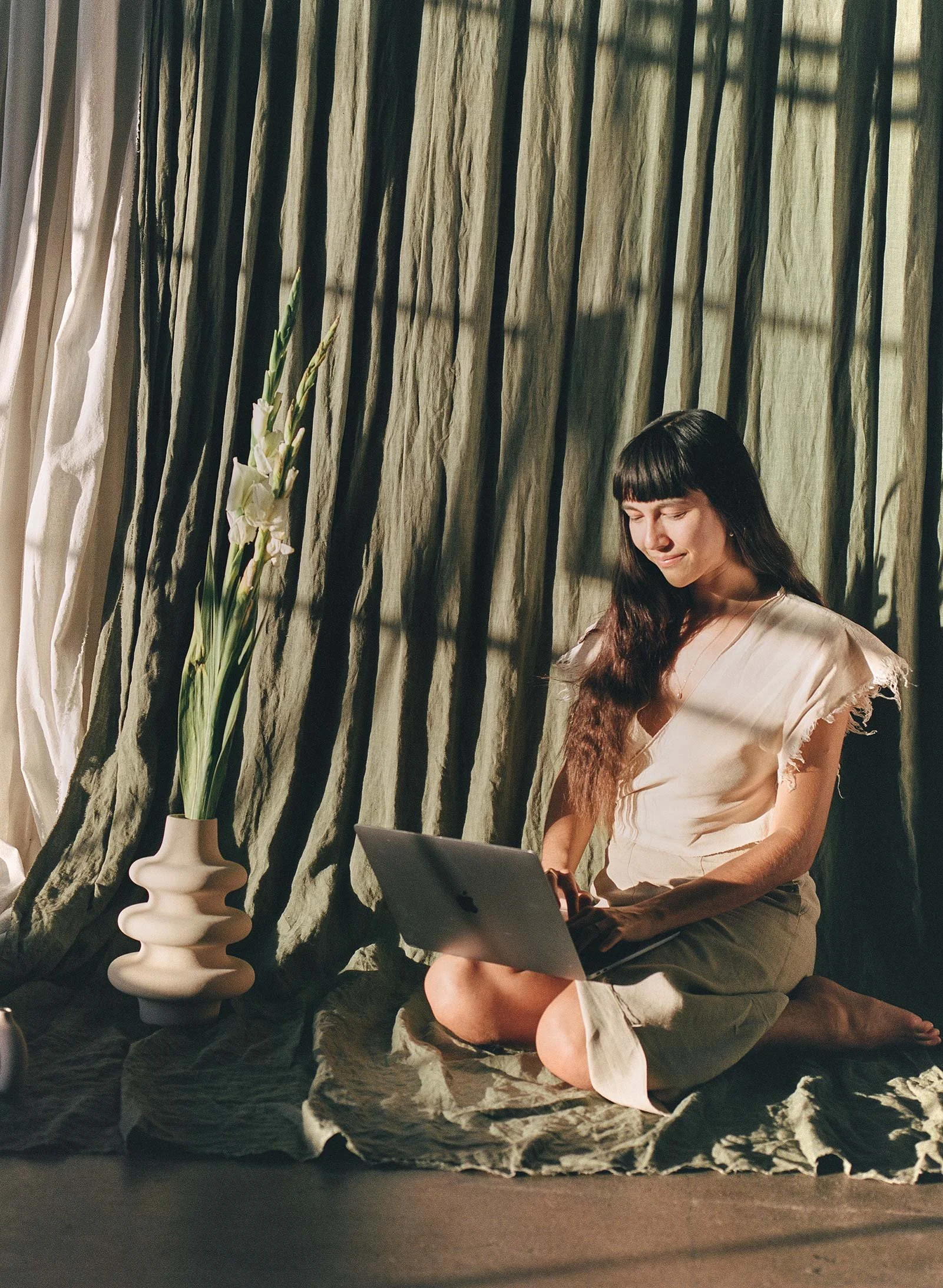 A woman with dark hair and bangs sitting on a rock-like surface, using a laptop. Behind her, floor-length curtains in shades of green and beige. To her left, a tall off-white vase with long-stemmed white flowers.