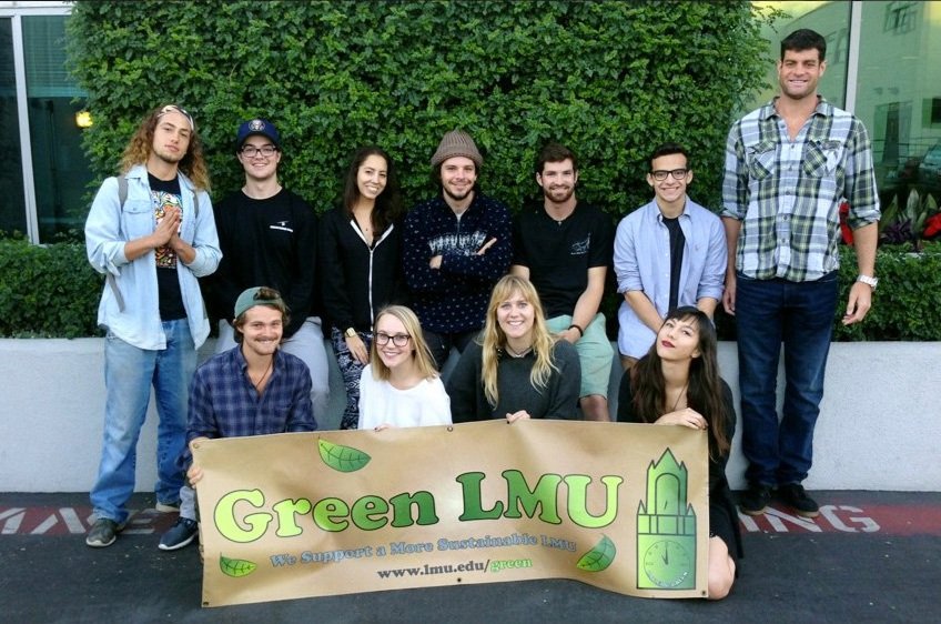 Group of ten young adults, some standing and some kneeling, holding a large beige banner that reads "Green LMU" supporting sustainable LMU, outdoors with green bushes and a glass building in the background.