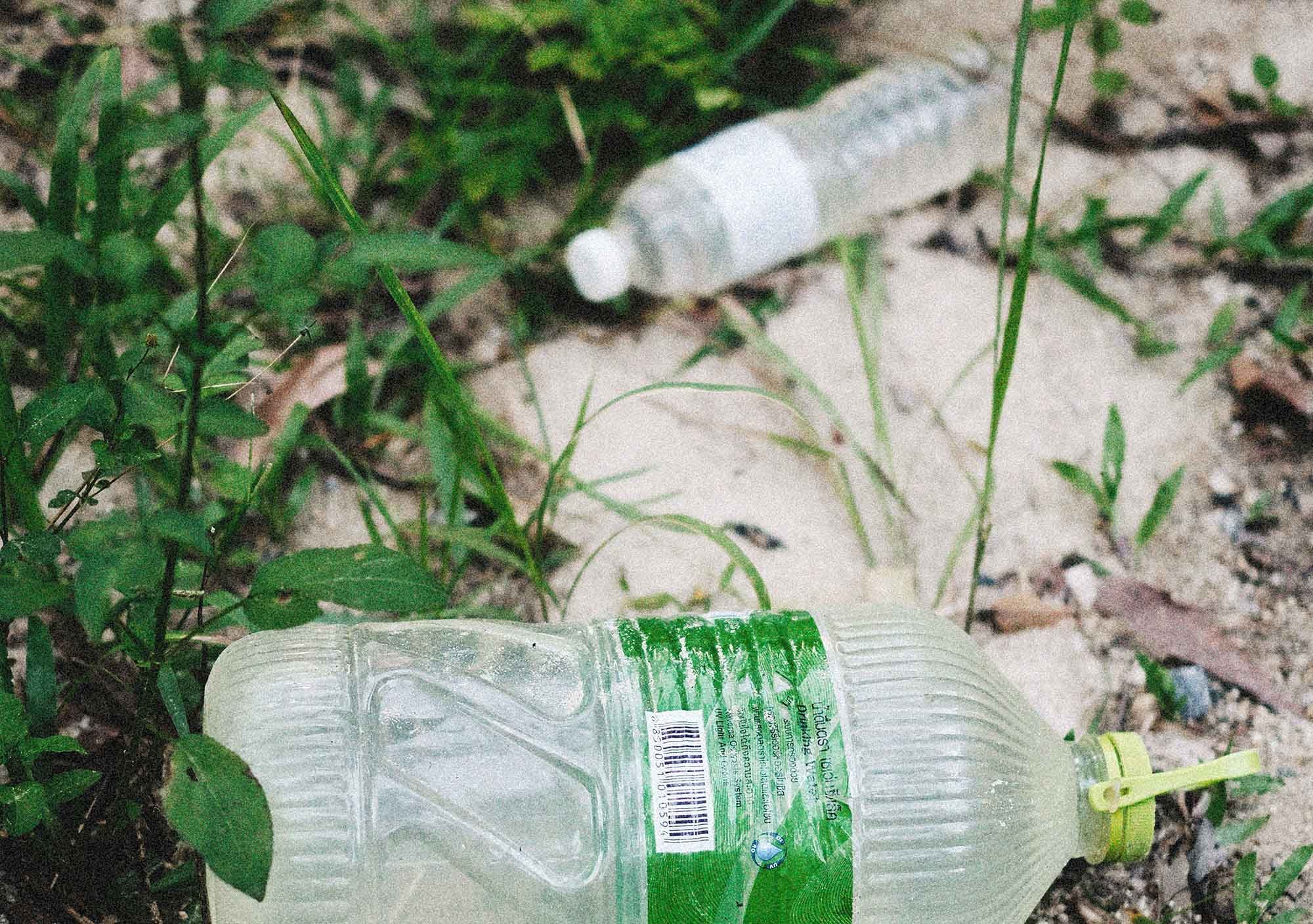 Discarded plastic water bottle with a green label lying on the ground among grass and dirt.