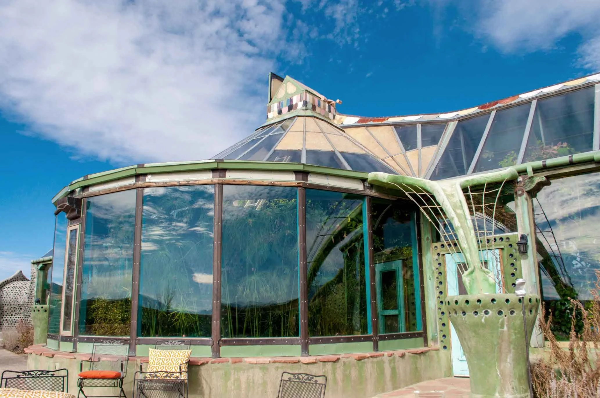 A greenhouse with large glass windows and a peaked roof, painted in shades of green and decorated with colorful panels and a small turret at the top, under a partly cloudy blue sky.