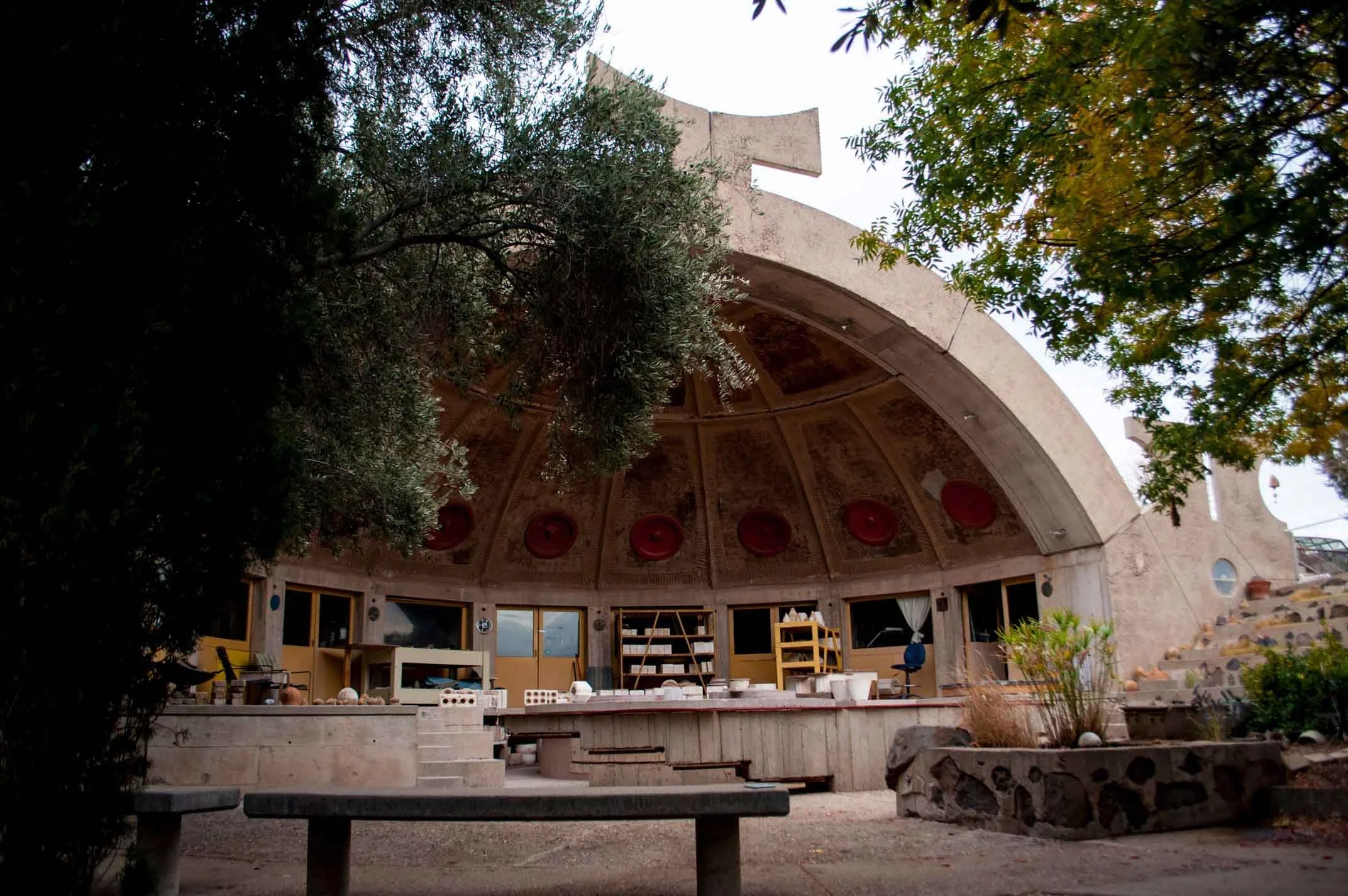Interior view of the Getty Villa with trees partially obscuring the view of the circular building's roof, which features artwork and architectural details, seen from a courtyard with chairs and furniture.
