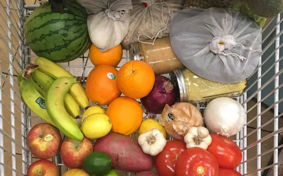 A shopping cart filled with various fruits and vegetables including watermelons, bananas, oranges, apples, a red onion, garlic, a white onion, tomatoes, a sweet potato, a jar of pickles, a package of what appears to be honey, and some produce wrapped in cloth bags.