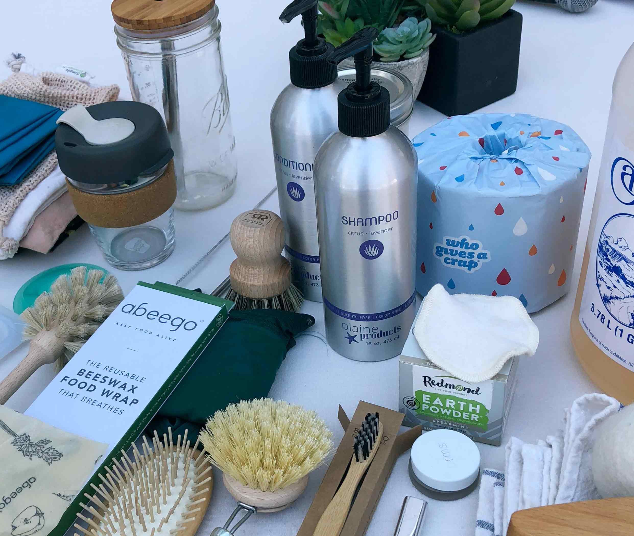 A collection of eco-friendly household items on a white surface, including spray bottles labeled shampoo and conditioner, a plant in a black pot, a jar, brushes, a bar of soap, and reusable cloth wipes with a blue and orange dotted pattern.