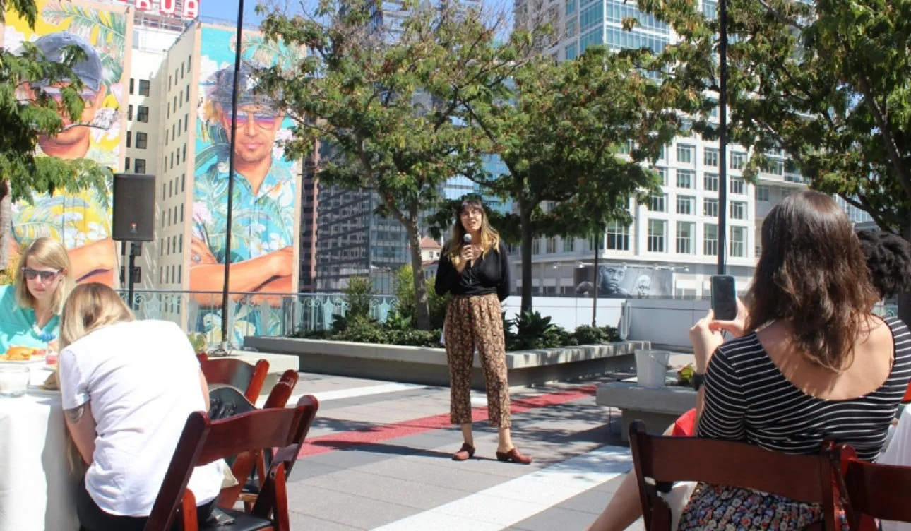 A woman performing a stand-up comedy routine on an outdoor rooftop terrace, with an audience seated at tables and a large mural of a man in sunglasses in the background.