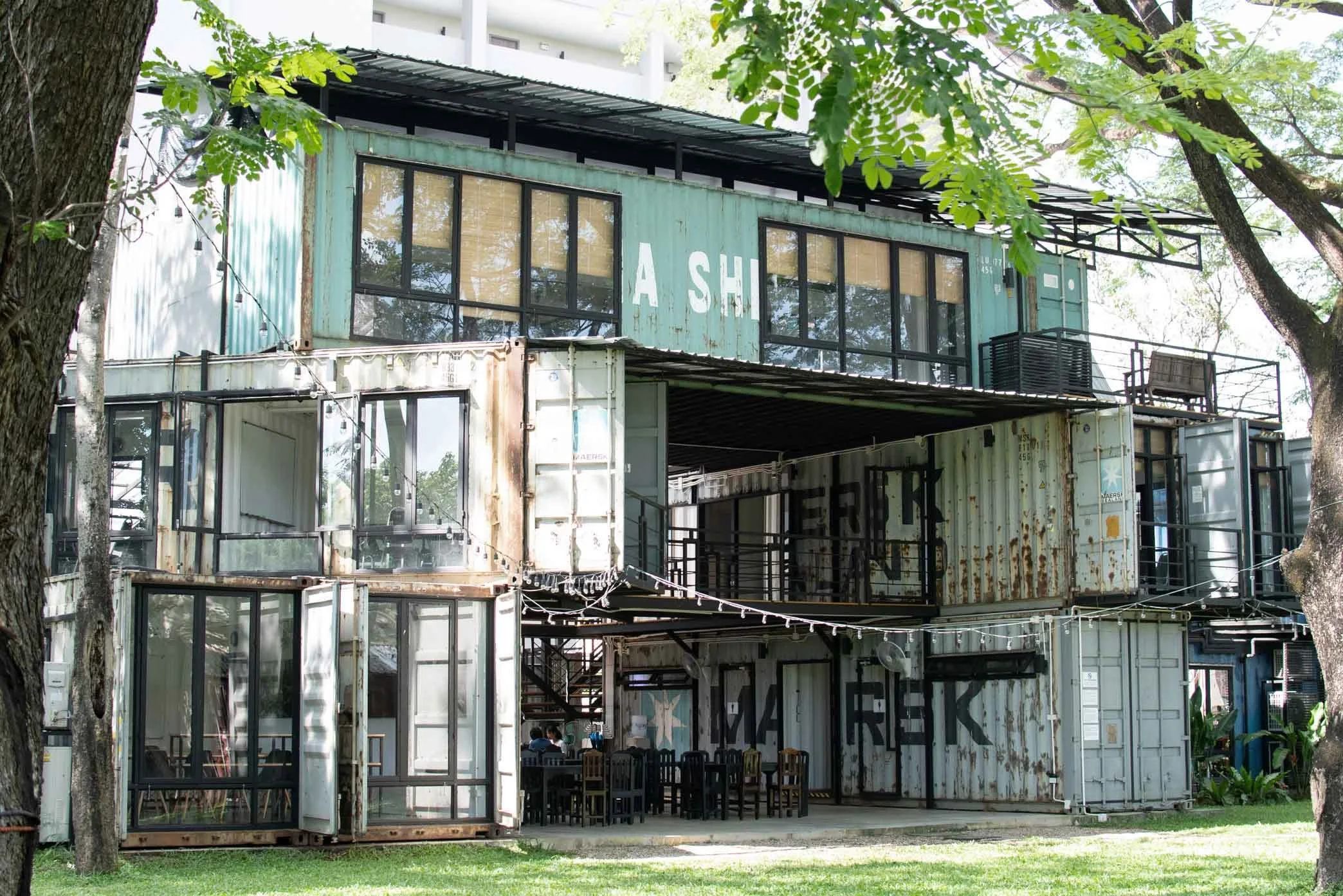 A building made of stacked shipping containers, including a green one with large windows and a partial sign that reads 'A SH.' The location is shaded by trees with green leaves, and there is a grassy area in front.