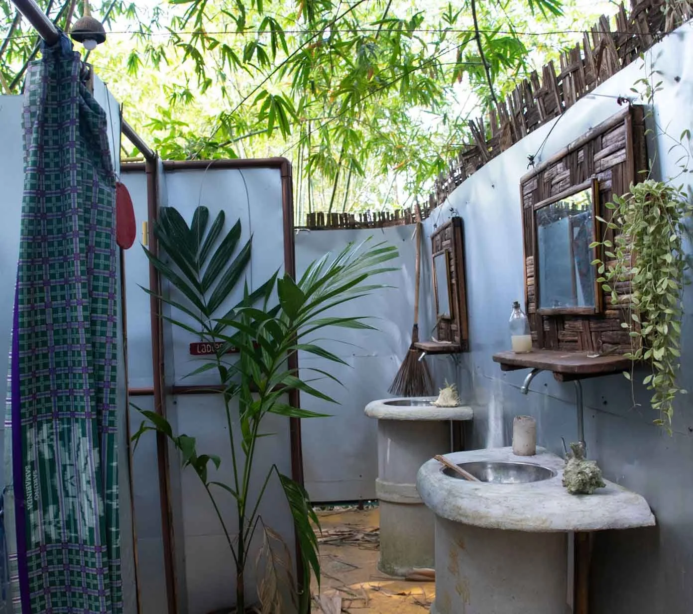 Outdoor rustic bathroom with two stone sinks, one with a soap dish, surrounded by green plants, bamboo, and wooden window frames, under a canopy of trees.