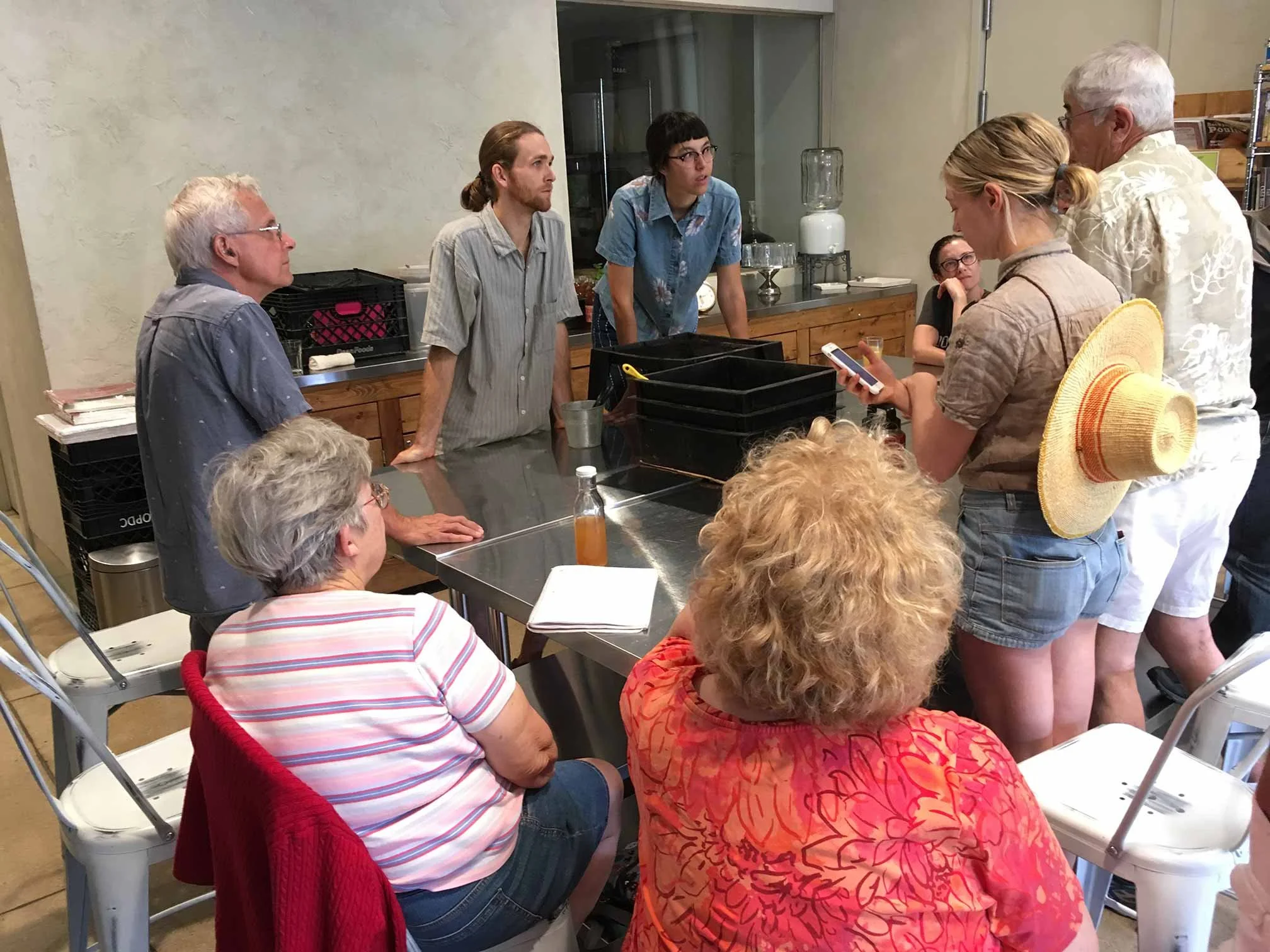 A group of people gathered around a stainless steel table, engaged in a discussion or presentation inside a room with beige walls. Some individuals are seated, while others stand, including a woman with a straw hat and a man with white hair. On the table are notebooks, a plastic bottle with an amber-colored liquid, and office supplies. The background features shelves, a water dispenser, and a window.
