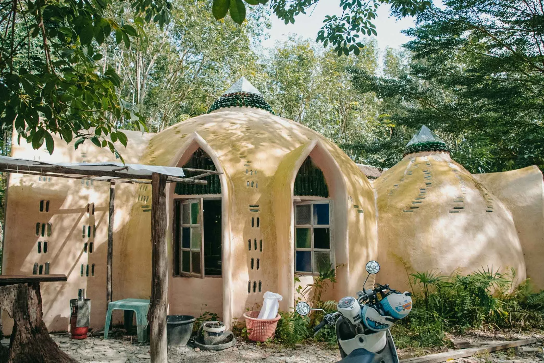 A house with a unique, organic shape, yellowish exterior walls, and small windows surrounded by lush green trees.