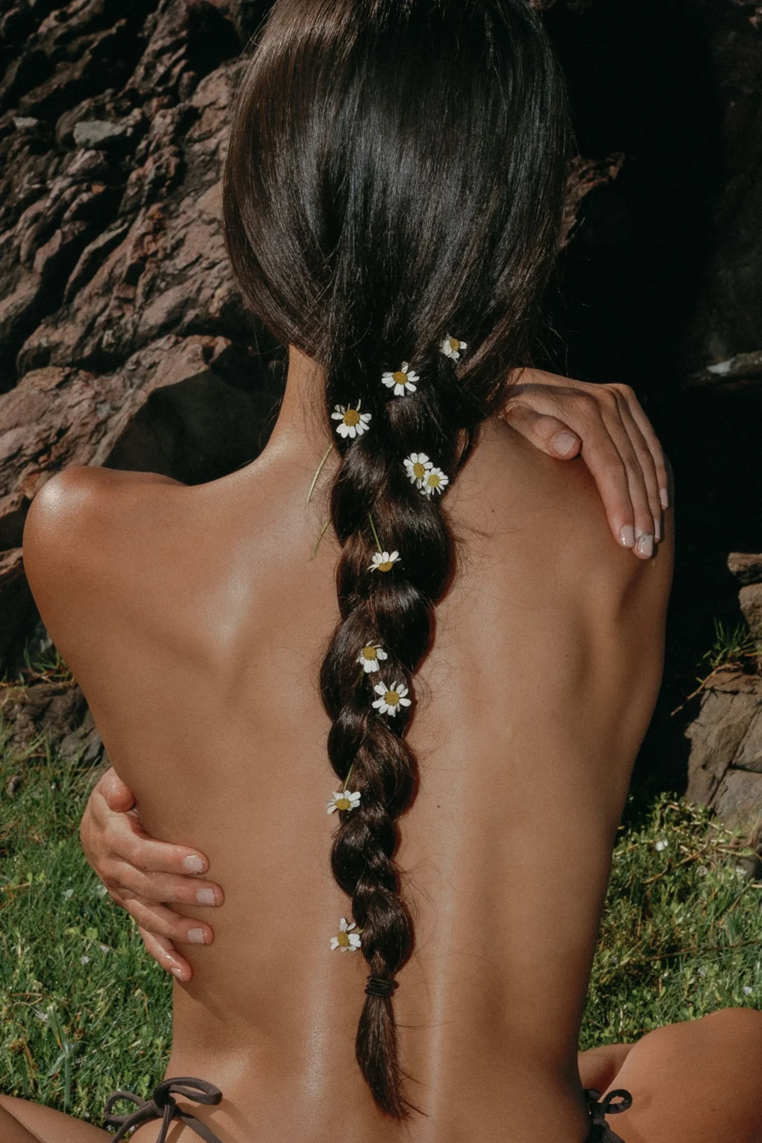A person with long, dark hair styled in a braid decorated with small white daisies with yellow centers, sitting outdoors with a rocky background and green grass visible.