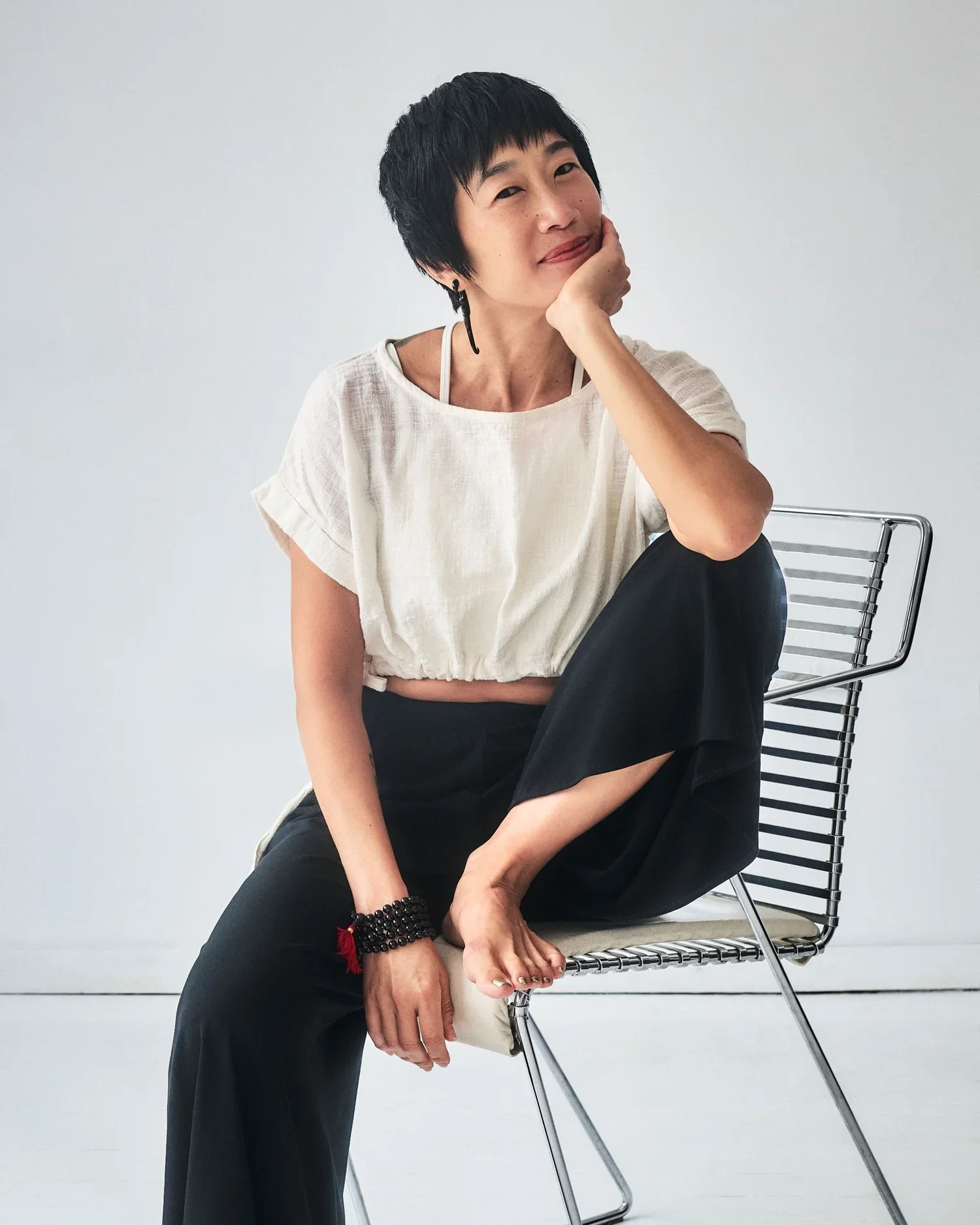 Female founder Jenny H Chao with short black hair, wearing a white loose top and black pants, sitting barefoot on a modern wire chair against a plain white background. She has a relaxed pose with one hand resting on her cheek.