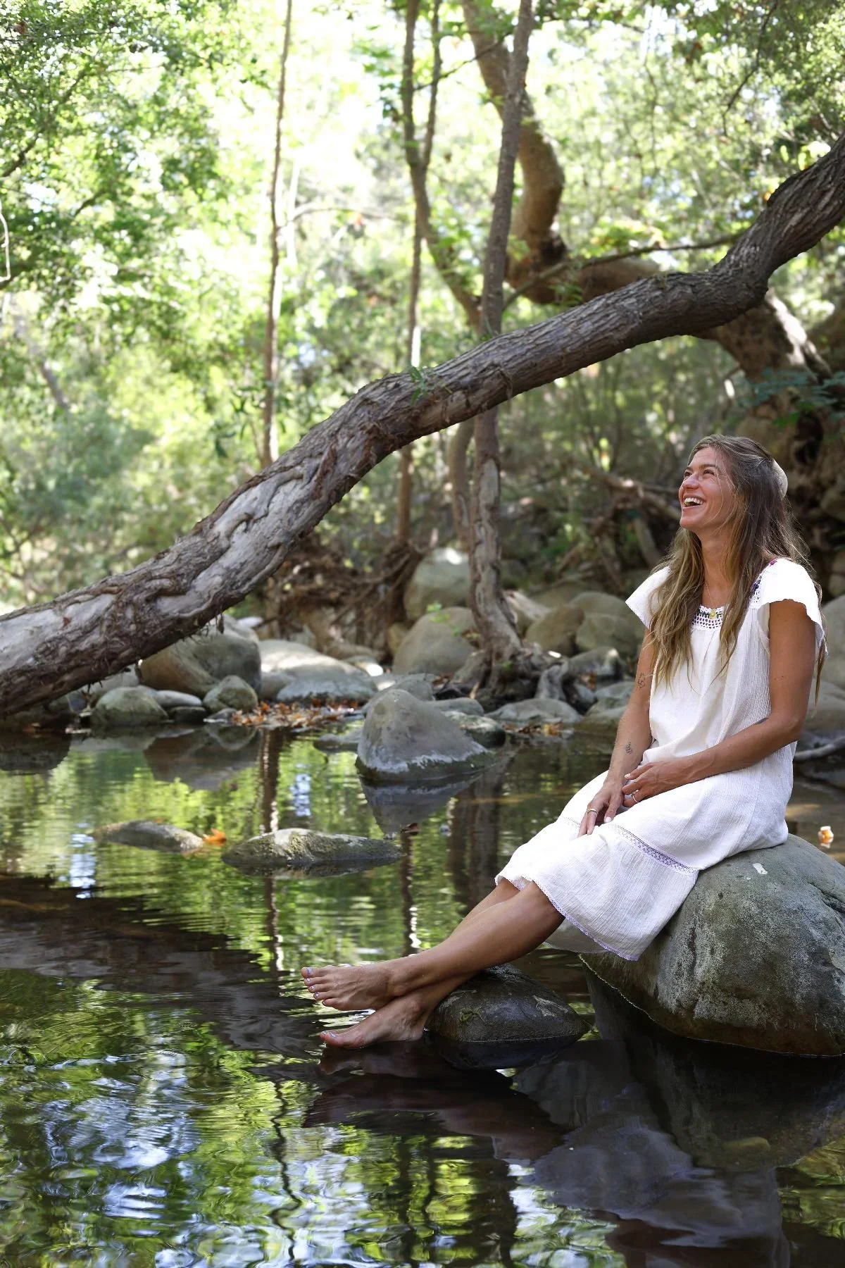 A woman in a white dress sitting on a rock by a creek in a forest, smiling and looking up, surrounded by trees and rocks with sunlight filtering through the leaves.