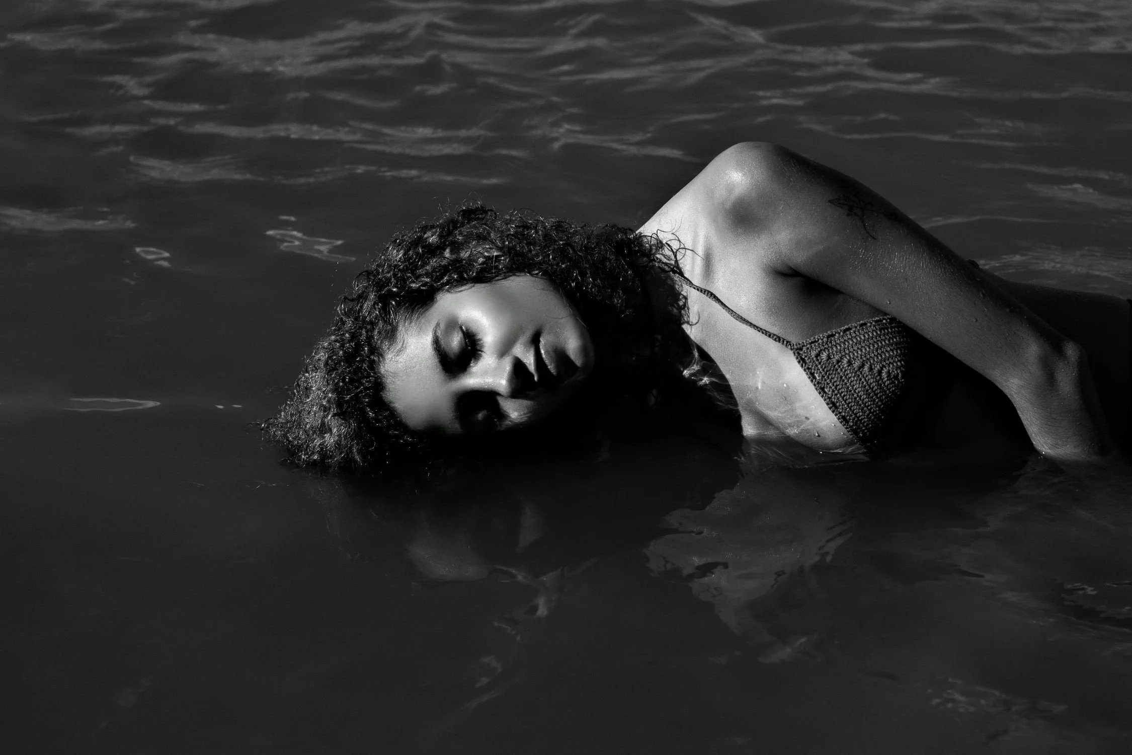 A woman with curly hair floating in the water with her eyes closed, wearing a swimsuit, in a black and white photograph.