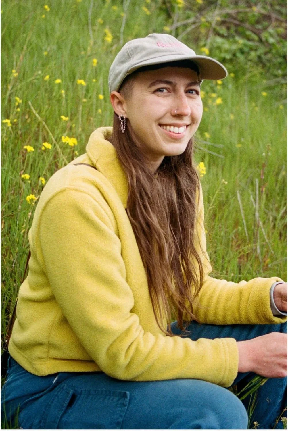 A young woman with long brown hair, wearing a beige cap, a yellow hoodie, and earrings, sitting in a field of tall grass and yellow flowers, smiling at the camera.