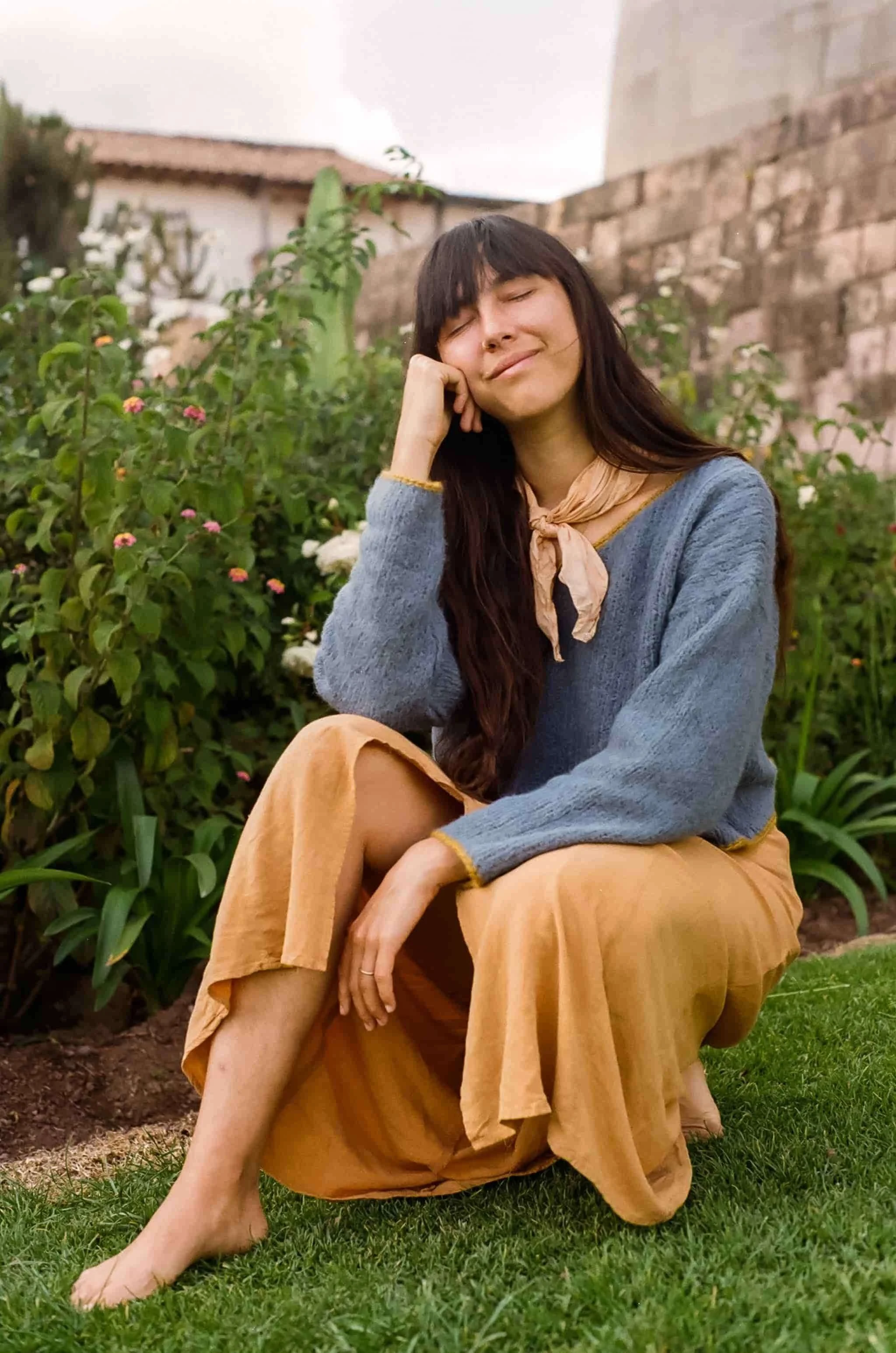 A woman with long dark hair, wearing a light scarf, a blue sweater, and a mustard-colored skirt, sitting with her eyes closed and smiling in a garden with green plants and a stone wall behind her.