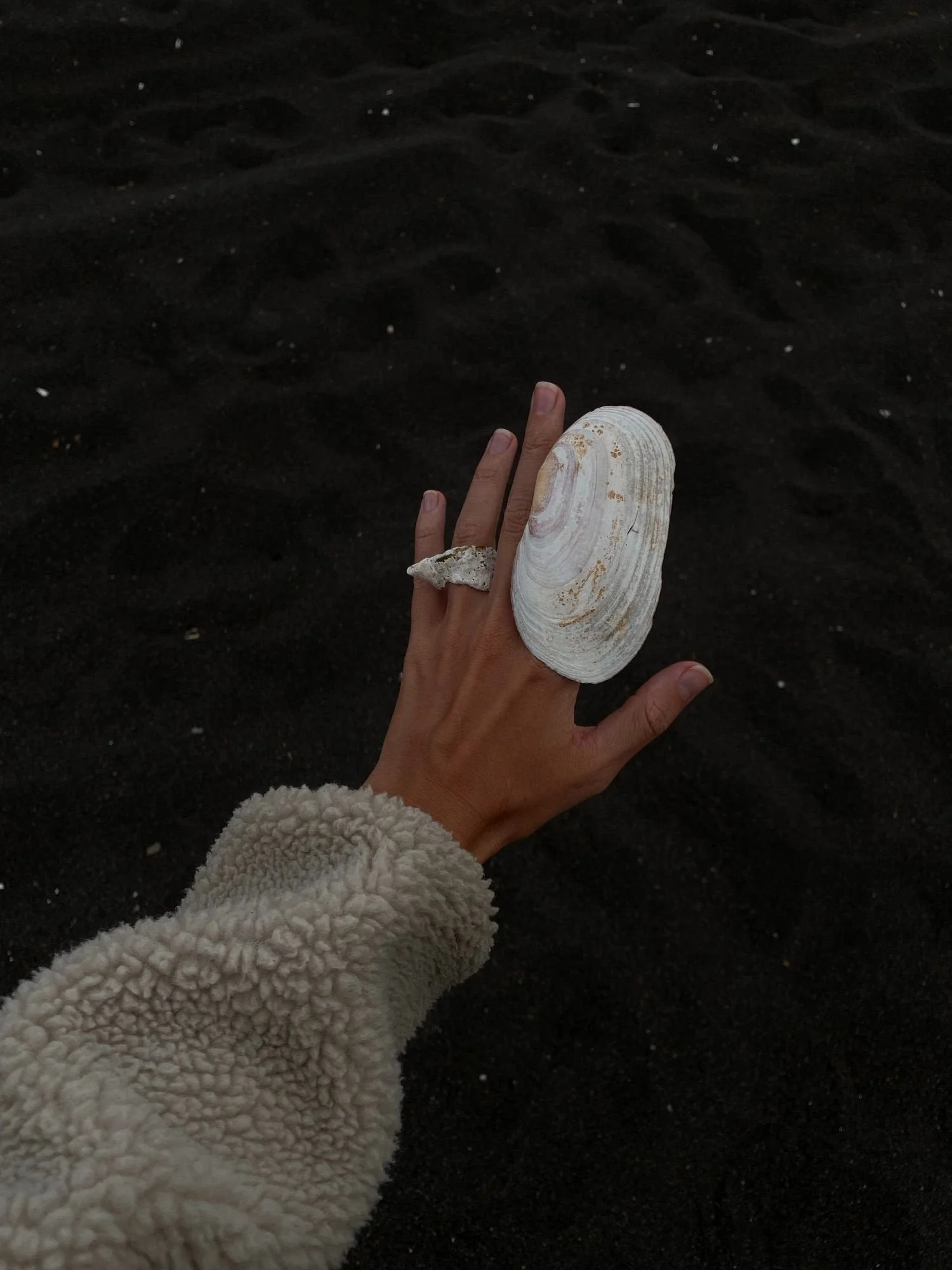 A dark image of a dark skinned woman putting her hand out, wearing shells as rings