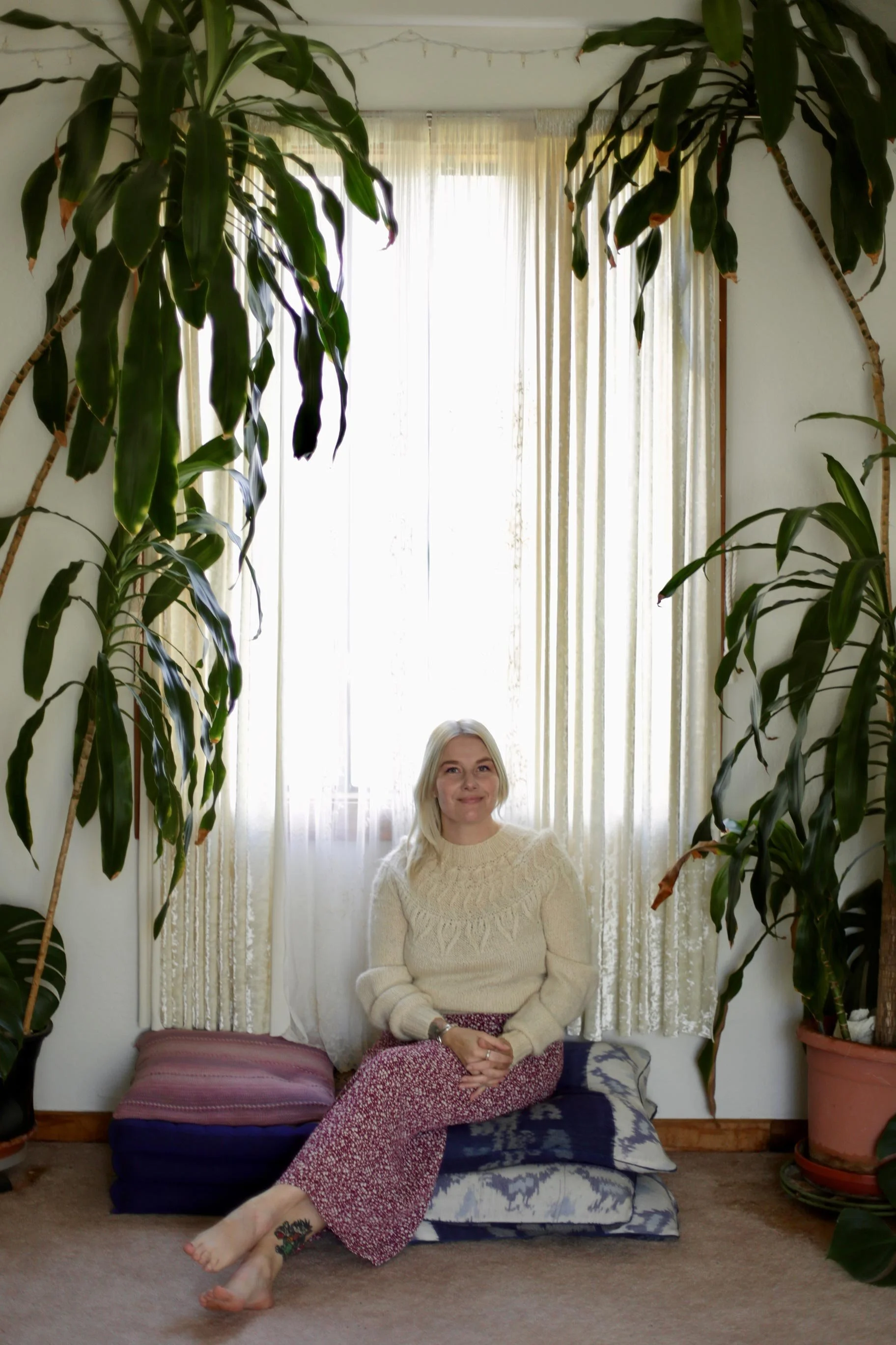 A woman sitting on cushions in front of a window with curtains, surrounded by large potted plants, smiling and looking at the camera.