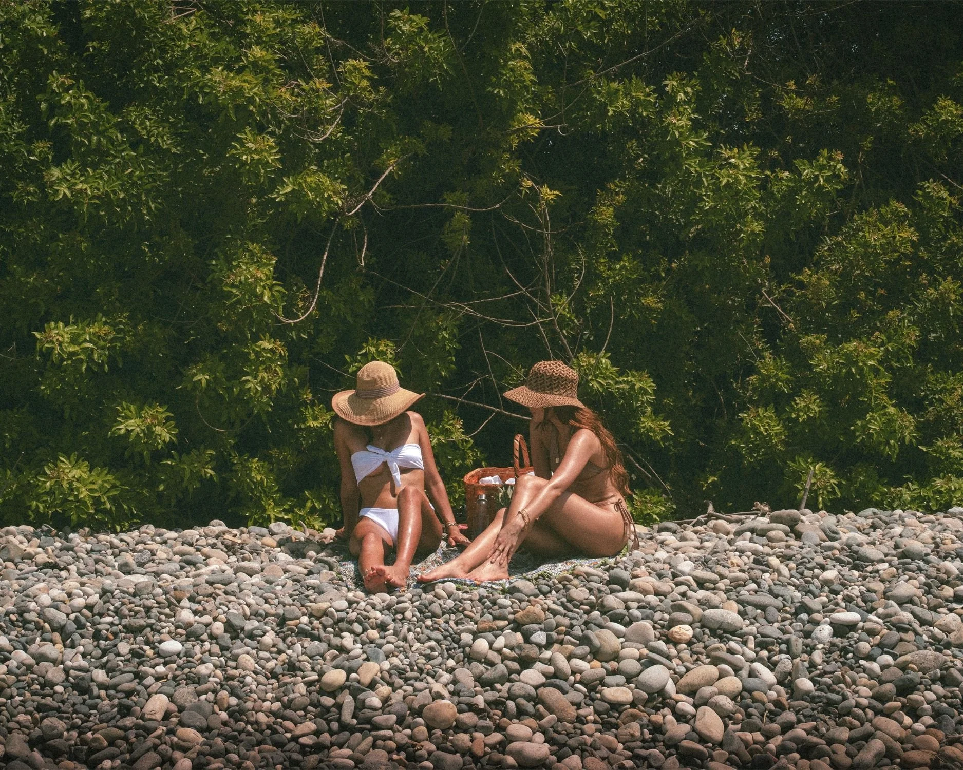Two women sitting on rocky beach with green foliage in the background, wearing swimwear and sun hats.