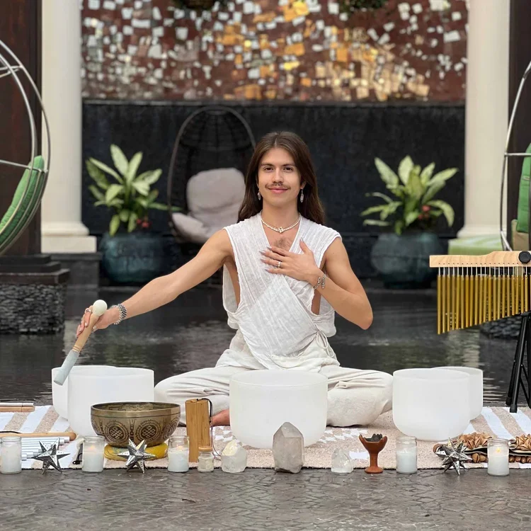 A woman with a mustache dressed in white sits cross-legged on the floor, surrounded by crystals, candles, and singing bowls, engaging in a spiritual or meditation session.