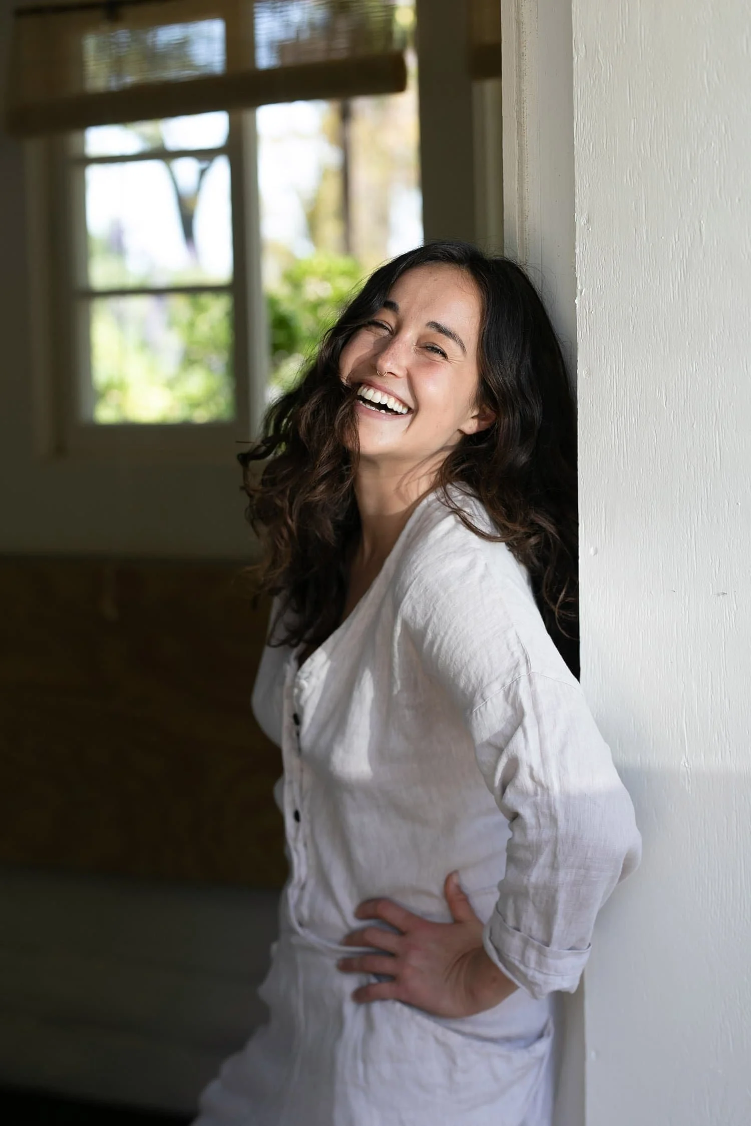 Female founder Erin Rose Ward with long, dark, curly hair leaning against a white wall indoors, smiling with her teeth showing, wearing a white linen shirt, with hands on her hips.
