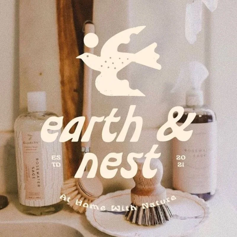 A personal care and cleaning products shelf featuring a bottle of exfoliating sugar scrub and a container of lotion, with a decorative wooden brush and a round woven mat, overlaid with a logo of a bird and the text 'earth & nest' and 'A Home With Nature'.