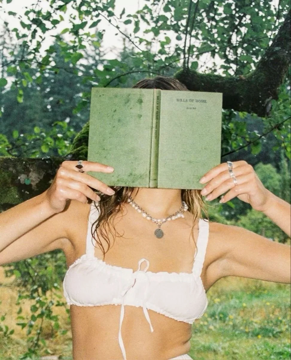 A woman wearing a white top and silver jewelry, holding a green book titled "Halls of Home" in front of her face, outdoors with leafy trees in the background.