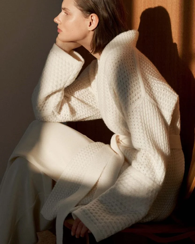A woman in a white textured sweater and cream-colored skirt sitting in profile against a dark background, with her head resting on her hand and soft lighting casting a shadow on the wall.