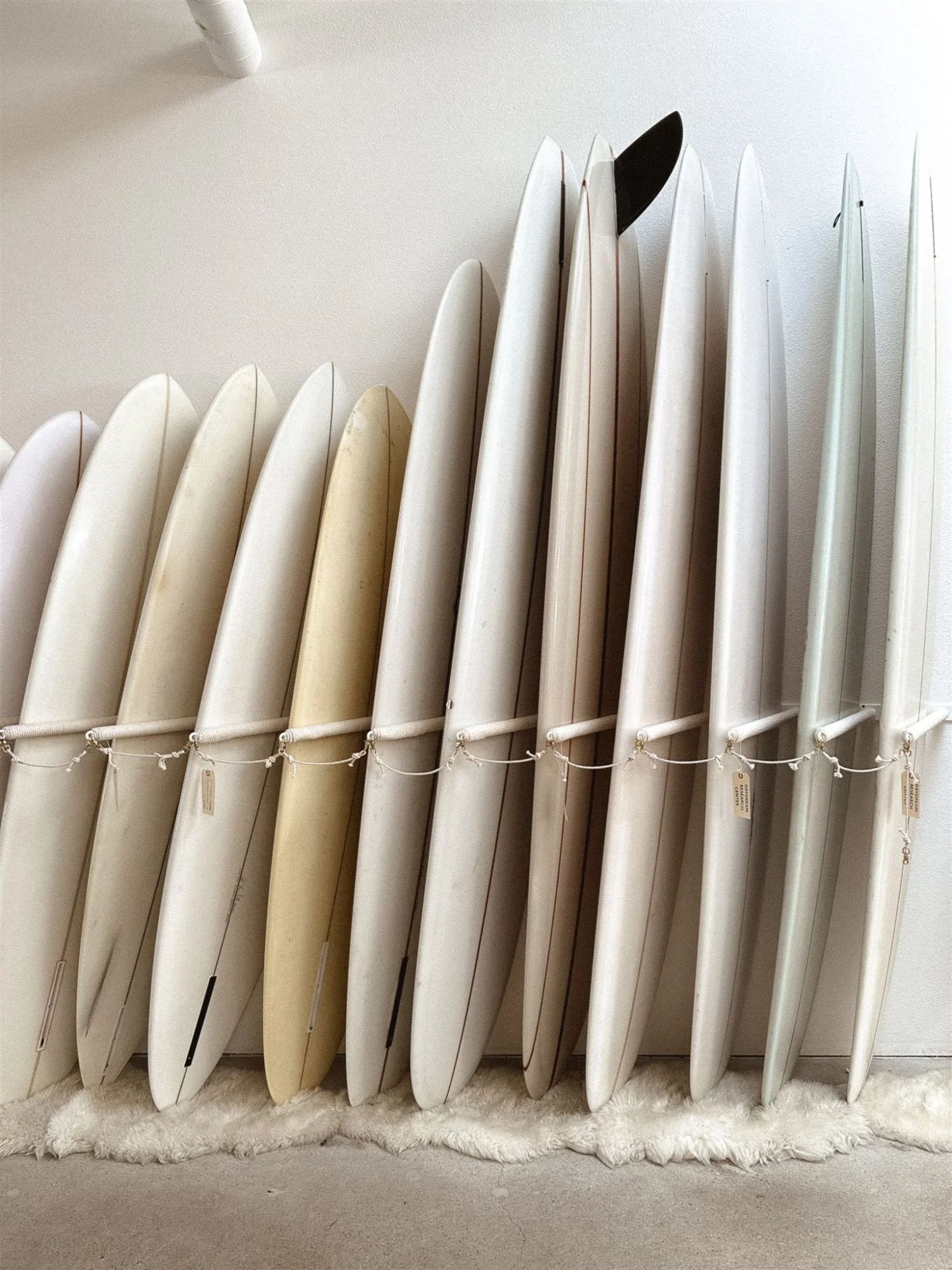 A row of surfboards standing upright on a white fluffy rug against a white wall, with some surfboards showing signs of wear and others appearing newer.