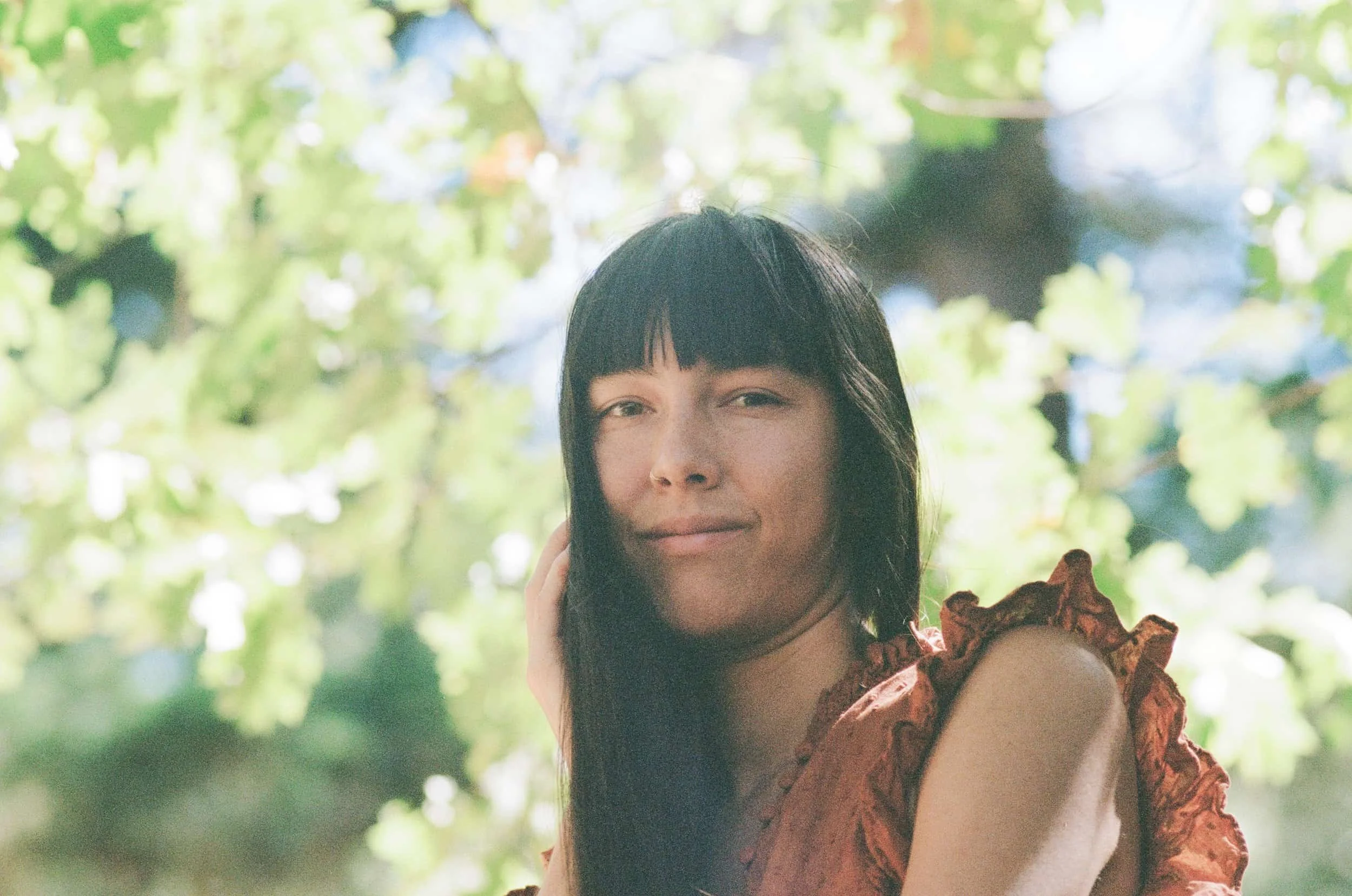 A woman with dark hair and bangs smiling outdoors, surrounded by green foliage, wearing a reddish-brown dress with ruffled sleeves.