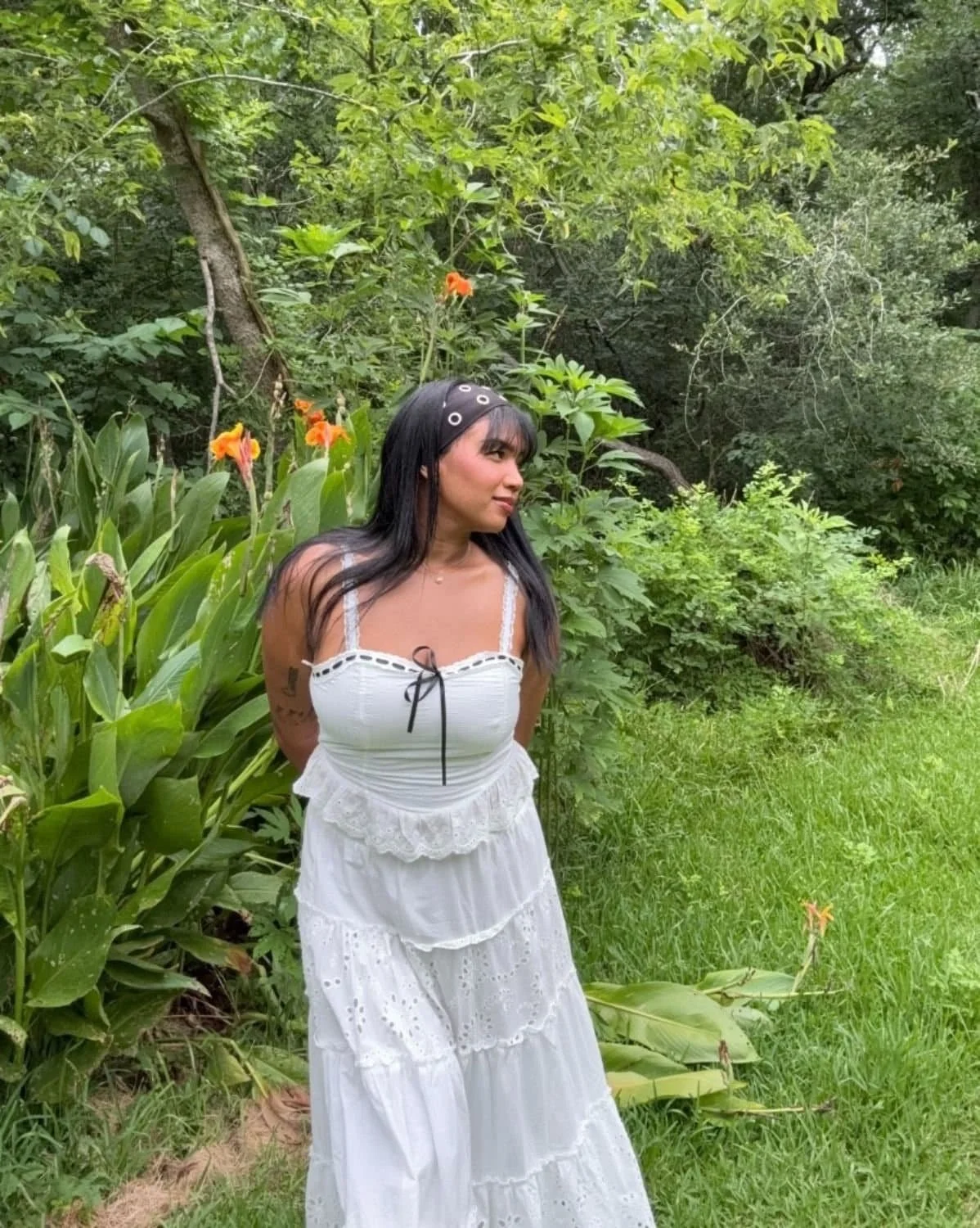 Female founder Lauren Janelle standing outdoors in a lush, green garden with orange flowers nearby, wearing a white eyelet dress and black headband with silver grommets.