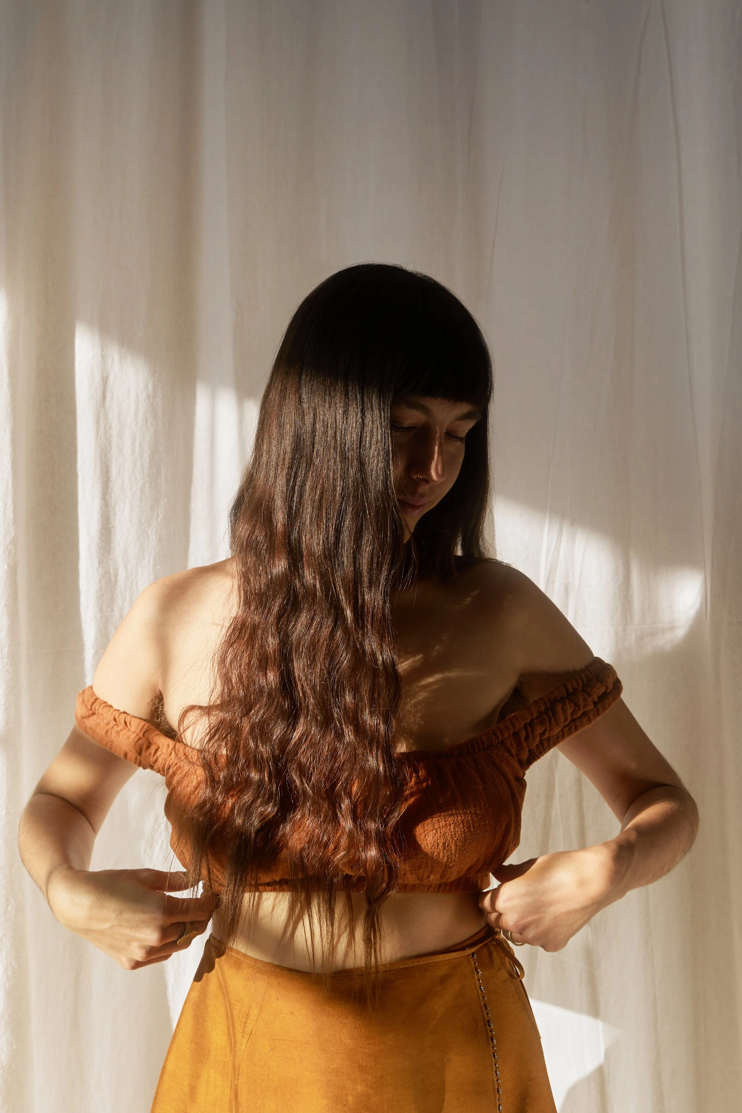 A woman with long wavy brown hair wearing a brown off-shoulder top and a high-waisted skirt, standing in front of a light-colored curtain, looking down with her eyes closed.