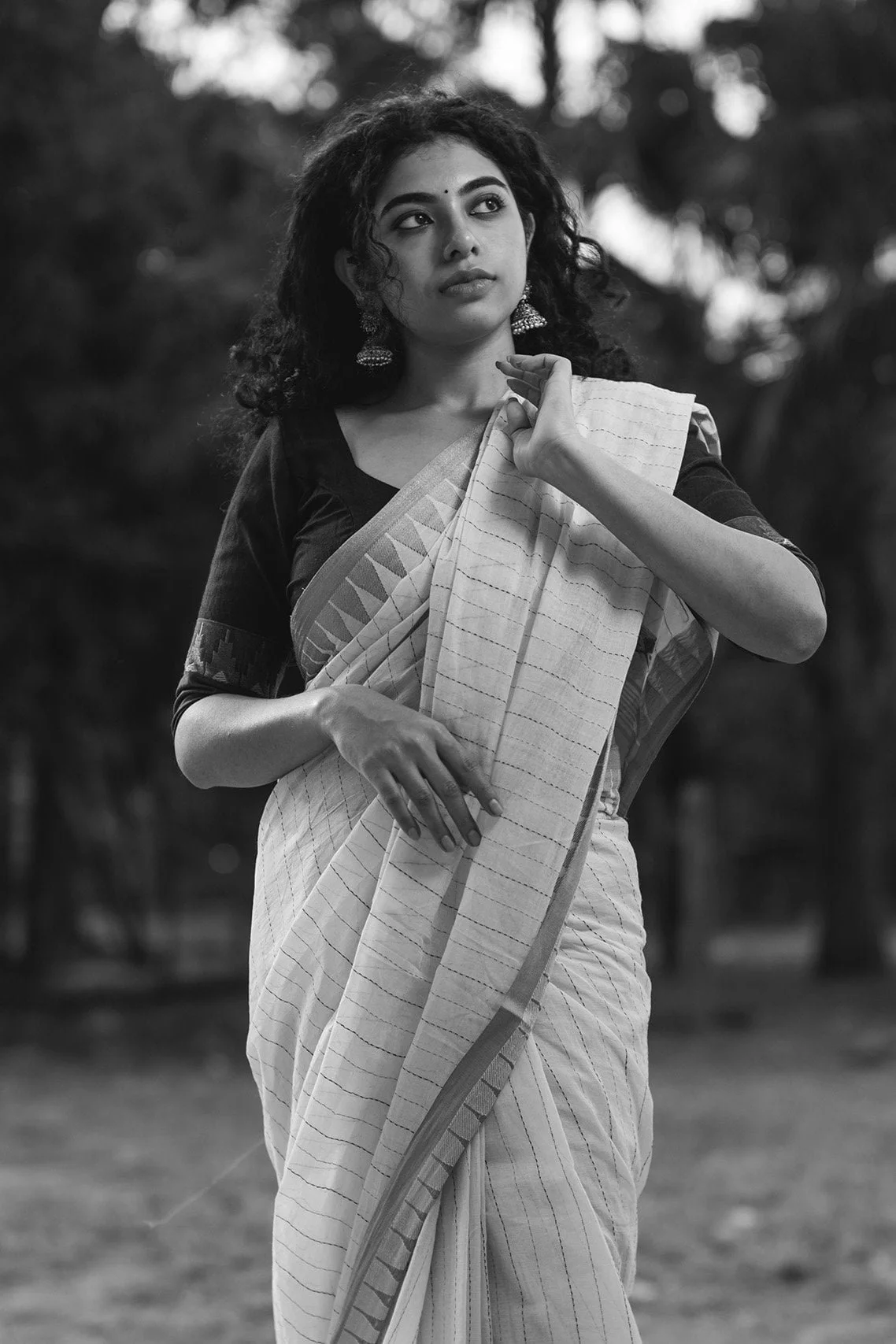 A woman in traditional Indian attire standing outdoors, holding a shawl draped over her shoulder, with trees in the background, black and white photograph for Tangachi.