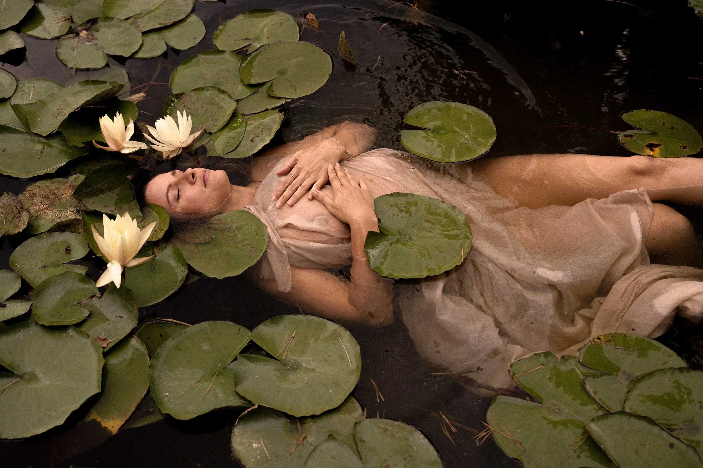 A woman is floating in water surrounded by lily pads and white water lilies.