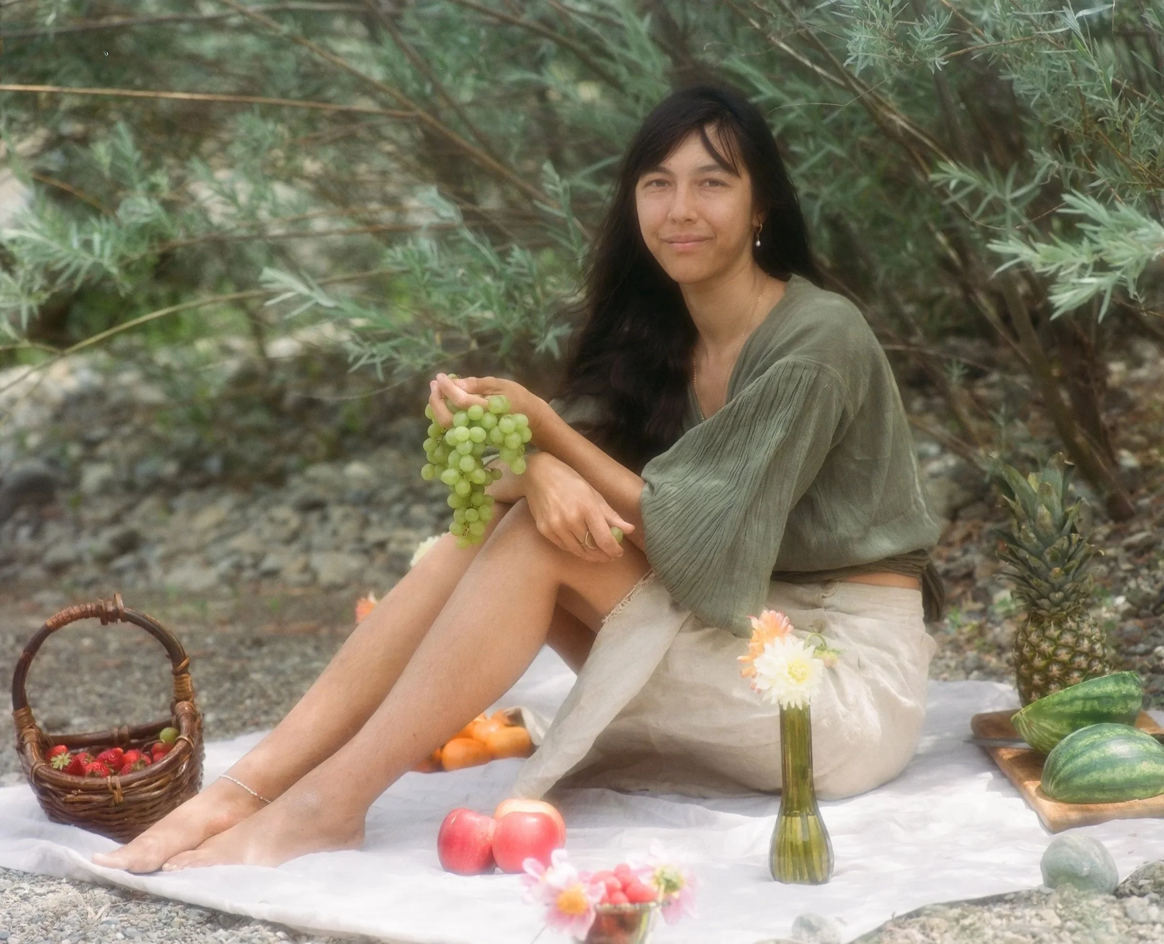A woman sitting outdoors among greenery, holding a bunch of green grapes, with a wicker basket of strawberries nearby, and a glass bottle with flowers, surrounded by various fruits and flowers on a white cloth.