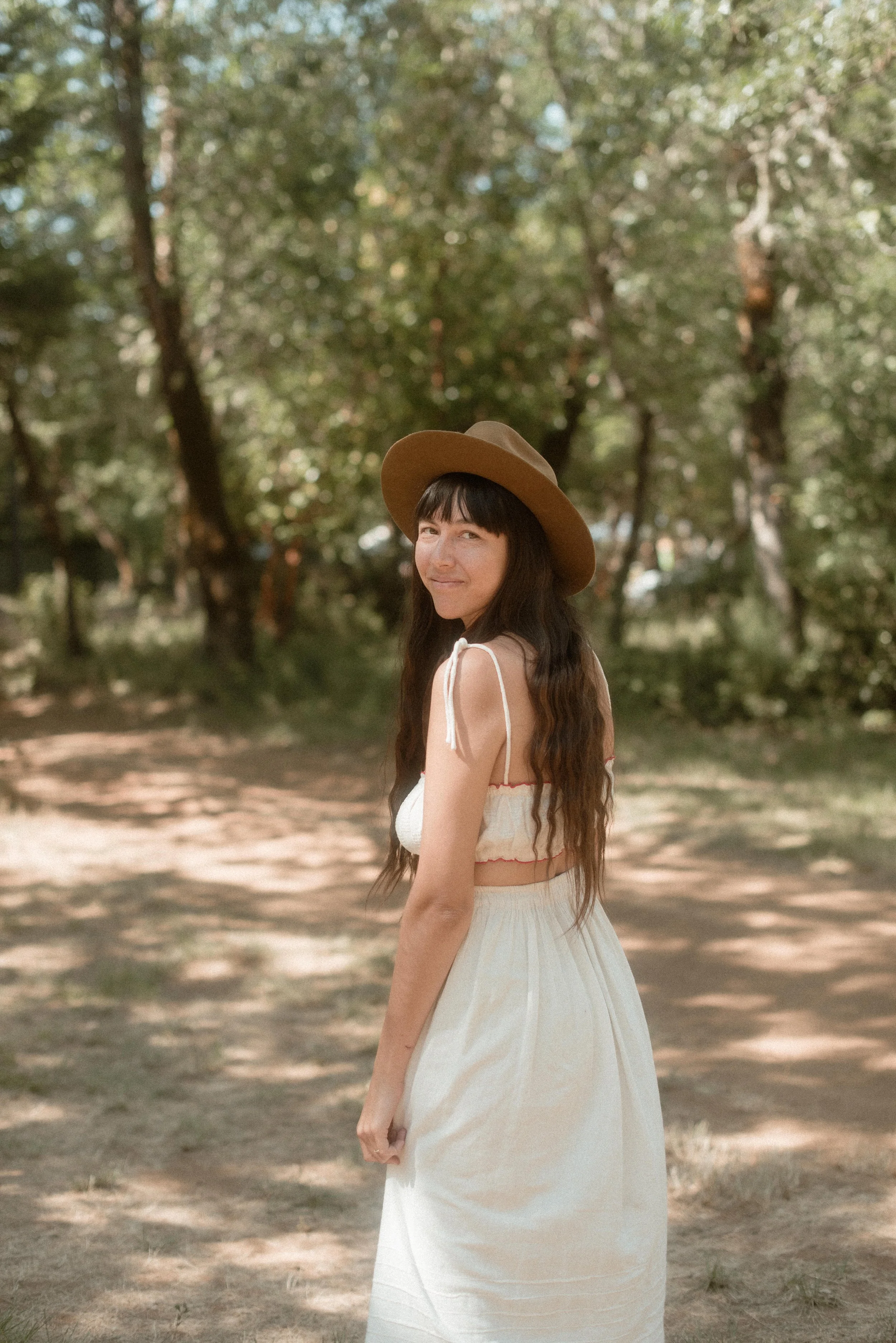 A woman wearing a wide-brimmed tan hat and a white dress with spaghetti straps standing outdoors in a wooded area with trees and sunlight filtering through the leaves.