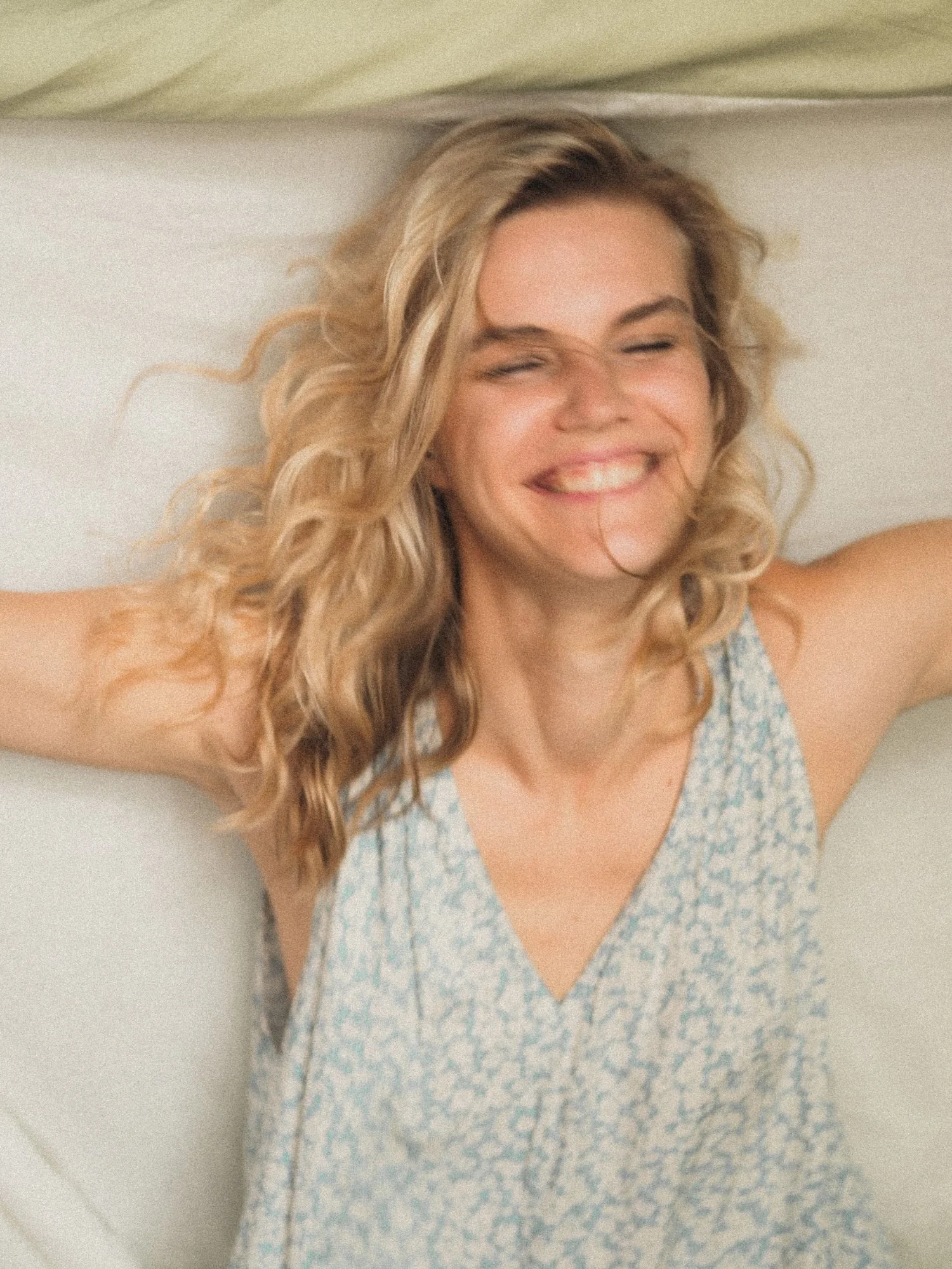 A woman with shoulder-length curly blonde hair smiling with closed eyes, wearing a sleeveless dress with a blue floral pattern.