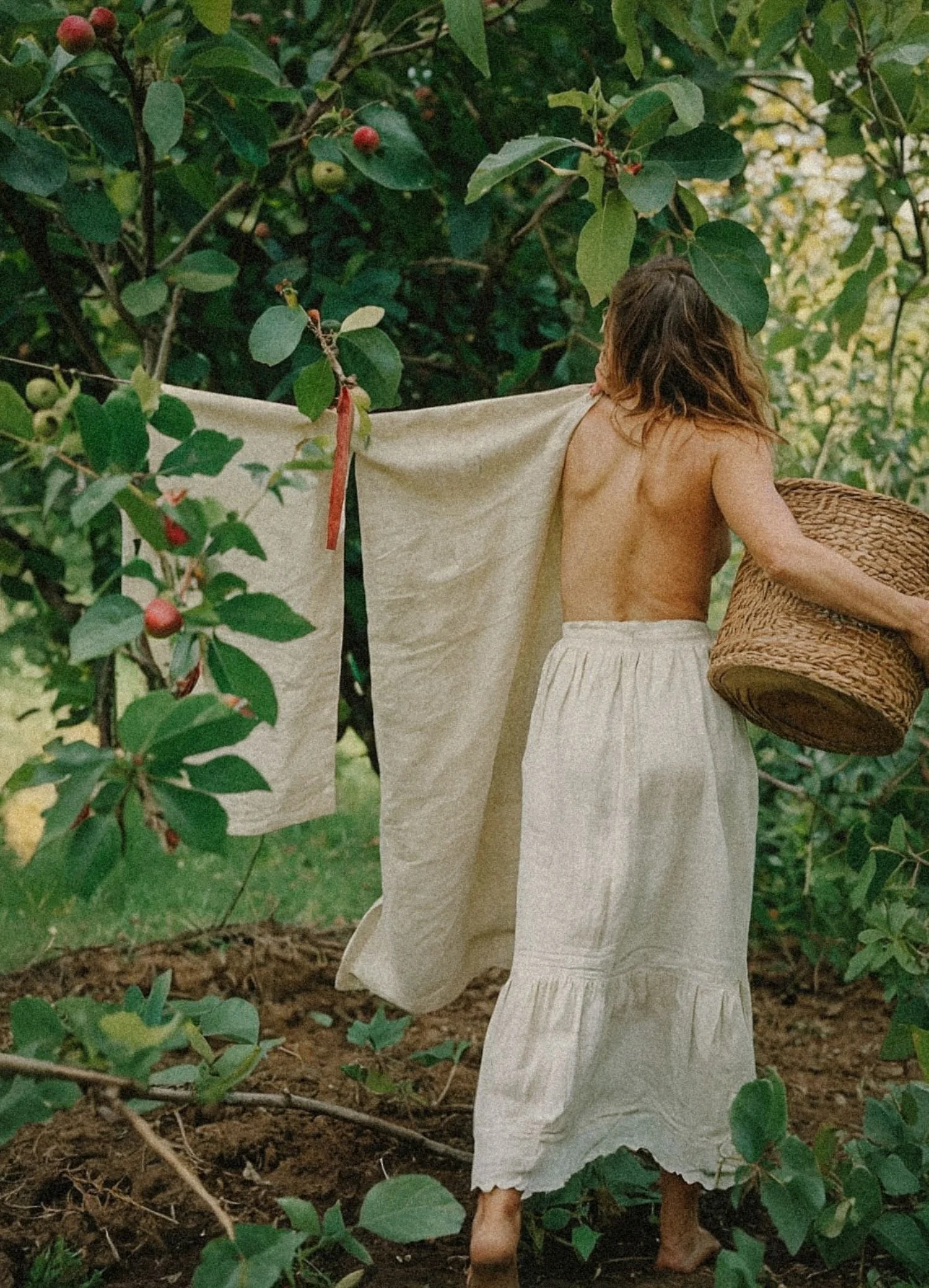 A person dressed in white, with long hair, standing among green leafy bushes, holding a large woven basket, and facing away from the camera.