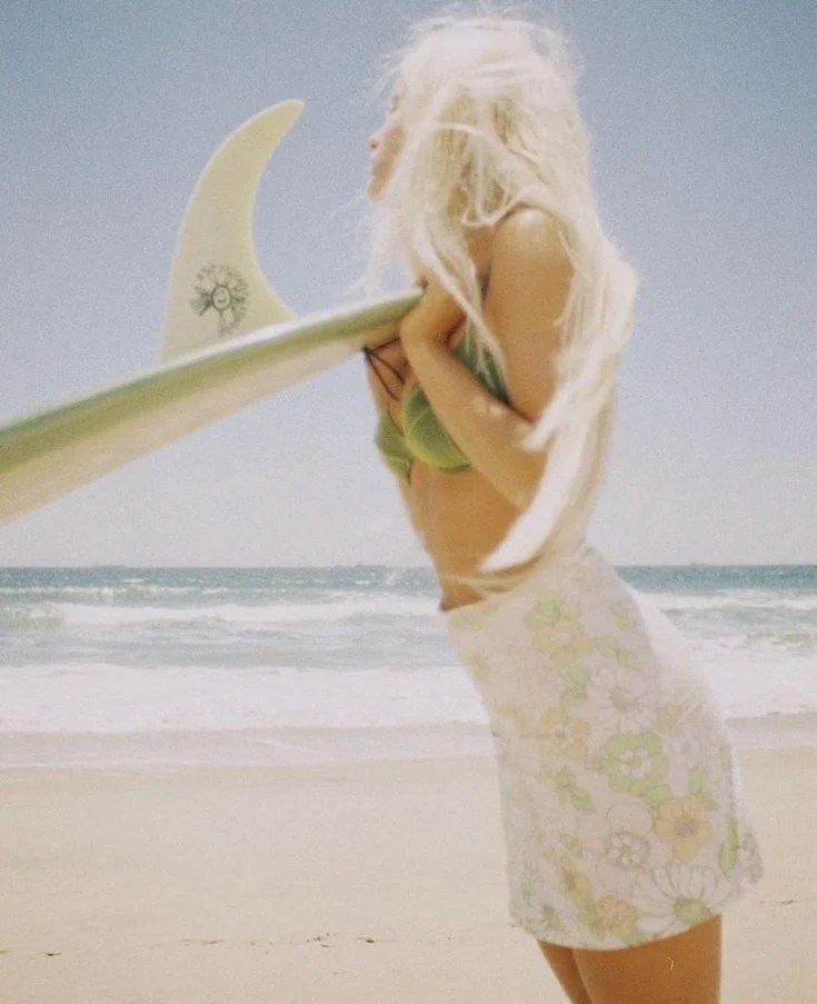 A woman with long blonde hair at the beach holding a surfboard under her arm, wearing a floral skirt and a green bikini top.