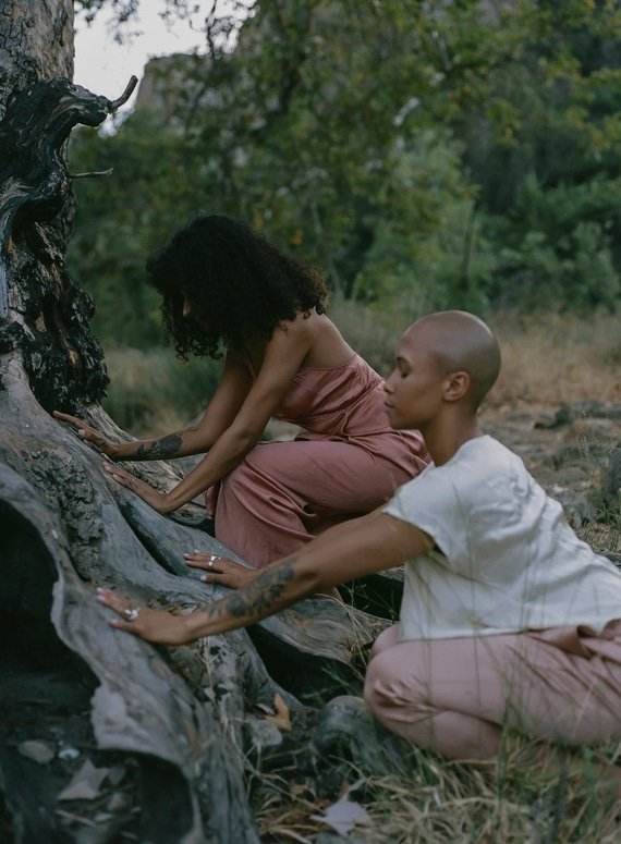 Two women with shaved heads and tattoos kneel and touch a large, fallen tree trunk outdoors in a natural setting for Of Beauty.
